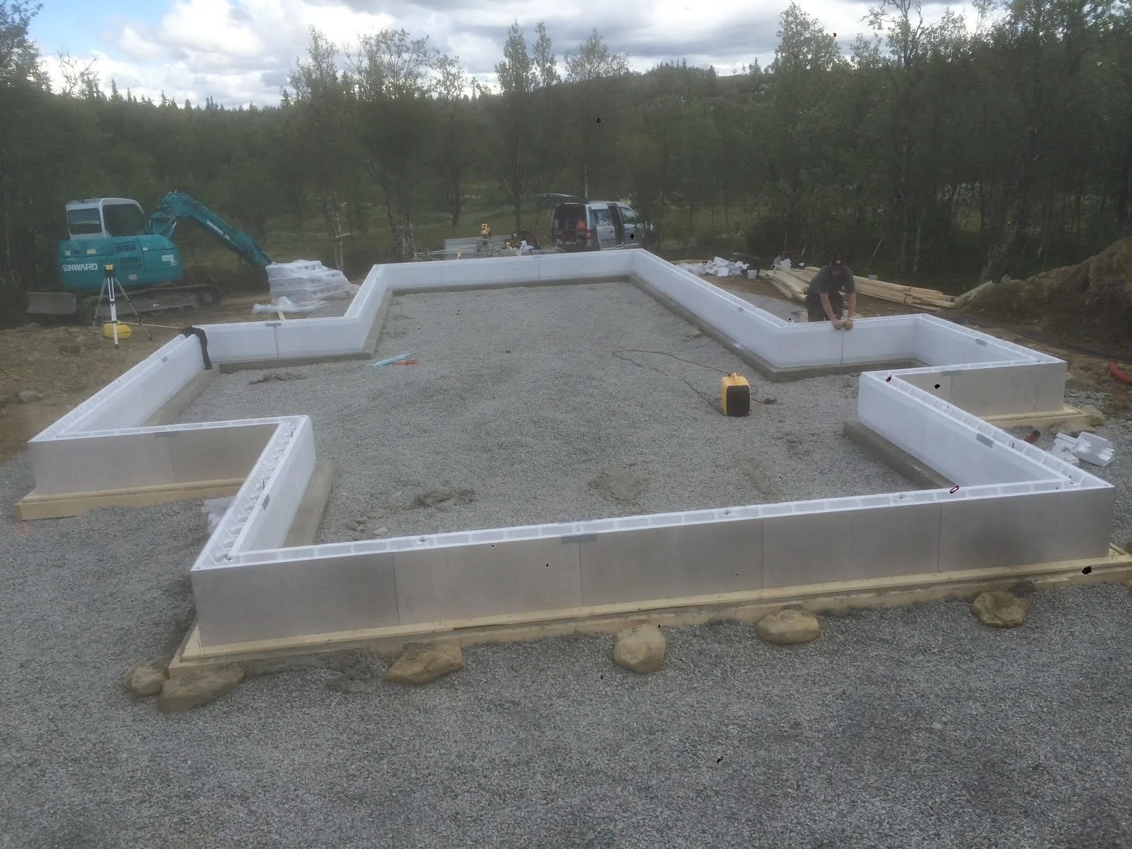 Construction site with an unfinished building foundation, workers installing insulation panels, a small excavator, and construction supplies nearby.