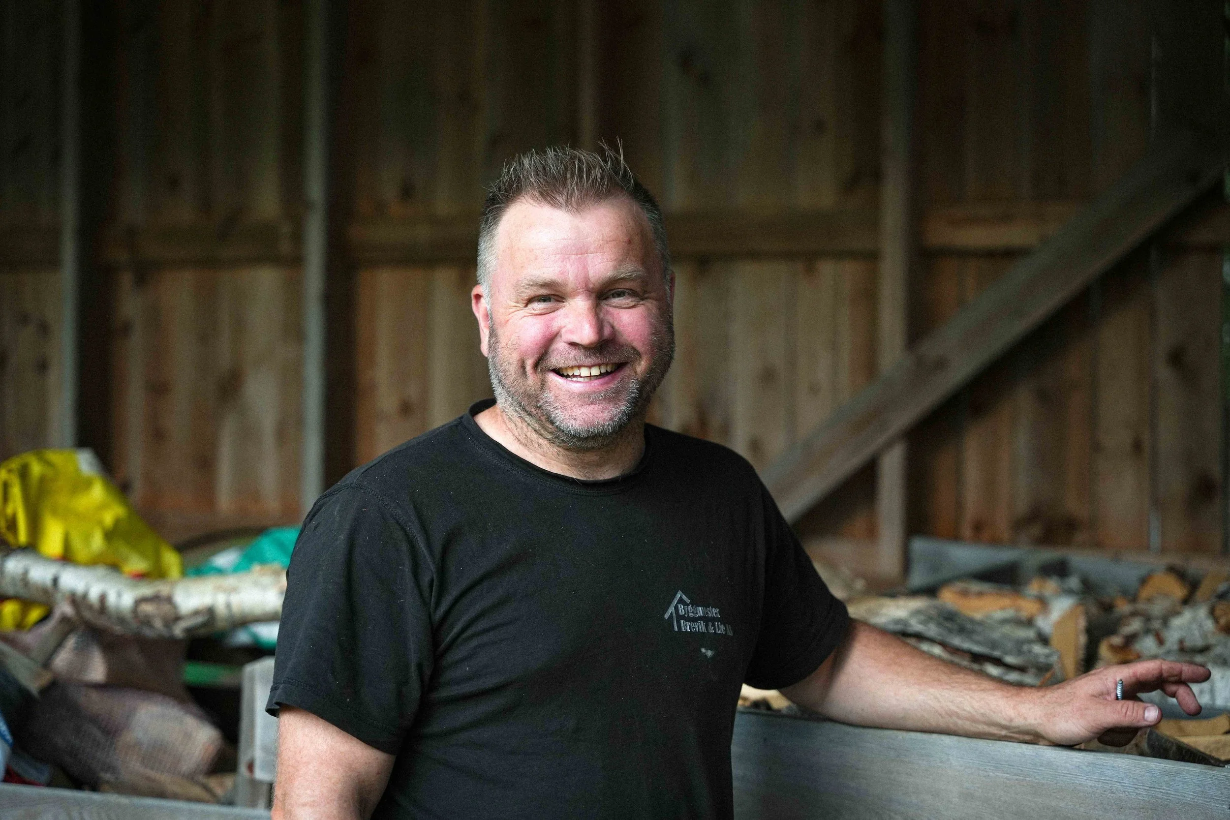 A smiling man with short hair and beard wearing a black shirt with a logo, standing in a woodworking or lumber storage area.