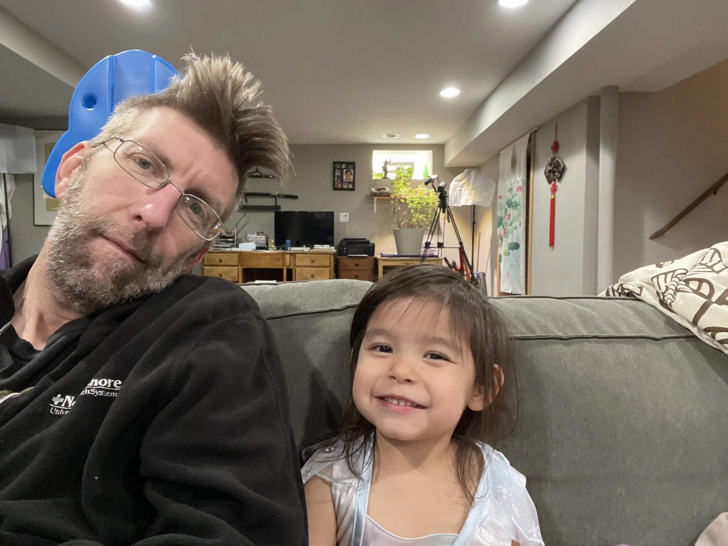 A man with glasses and a beard sitting on a gray couch next to a young girl with dark hair and a white dress, smiling in a living room with a television, camera tripod, and houseplants in the background.