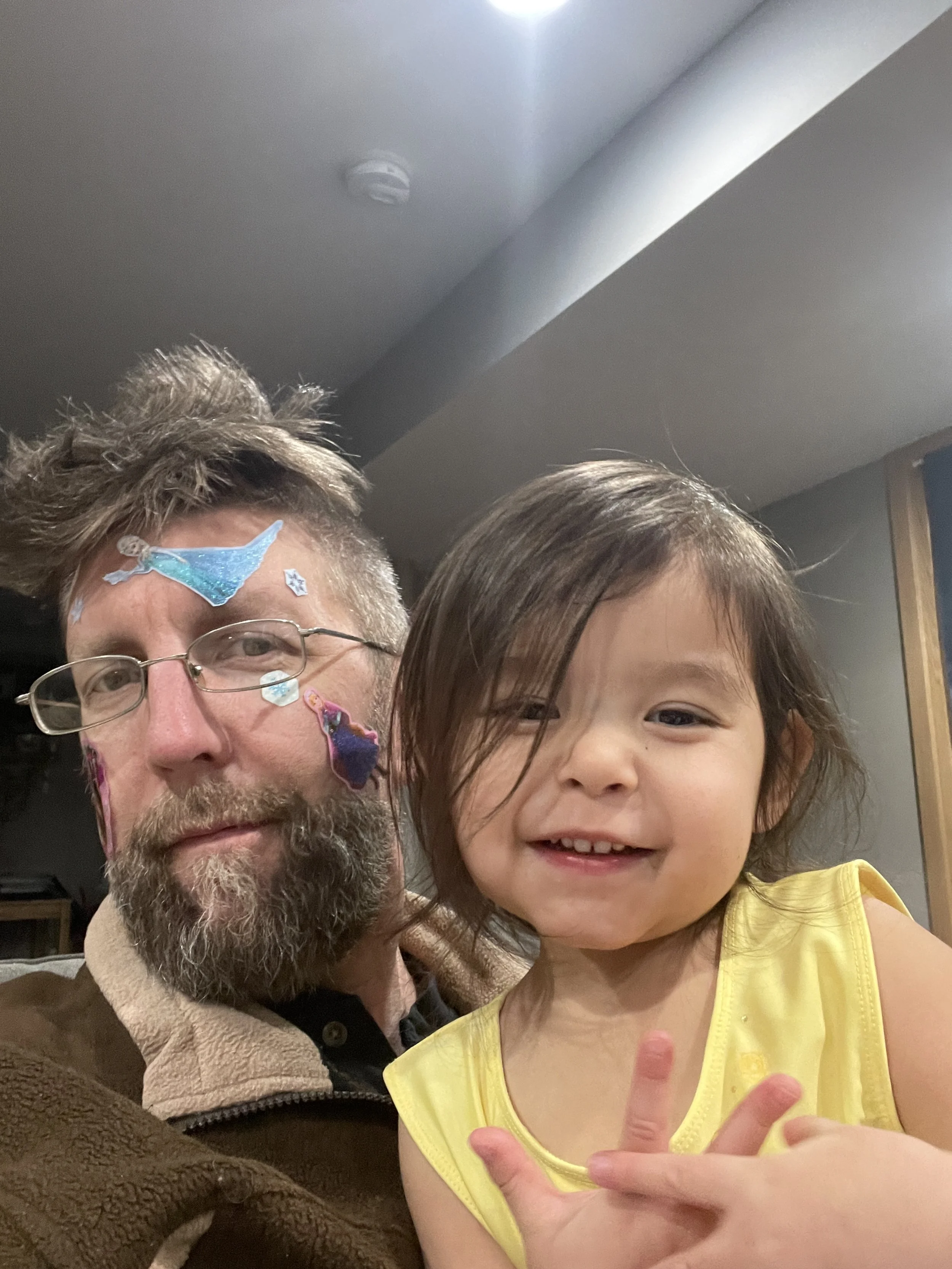 A man with glasses and a beard, and a young girl, both smiling. The man has colorful stickers on his face and hair, and the girl is wearing a yellow top, making a peace sign with her hand.