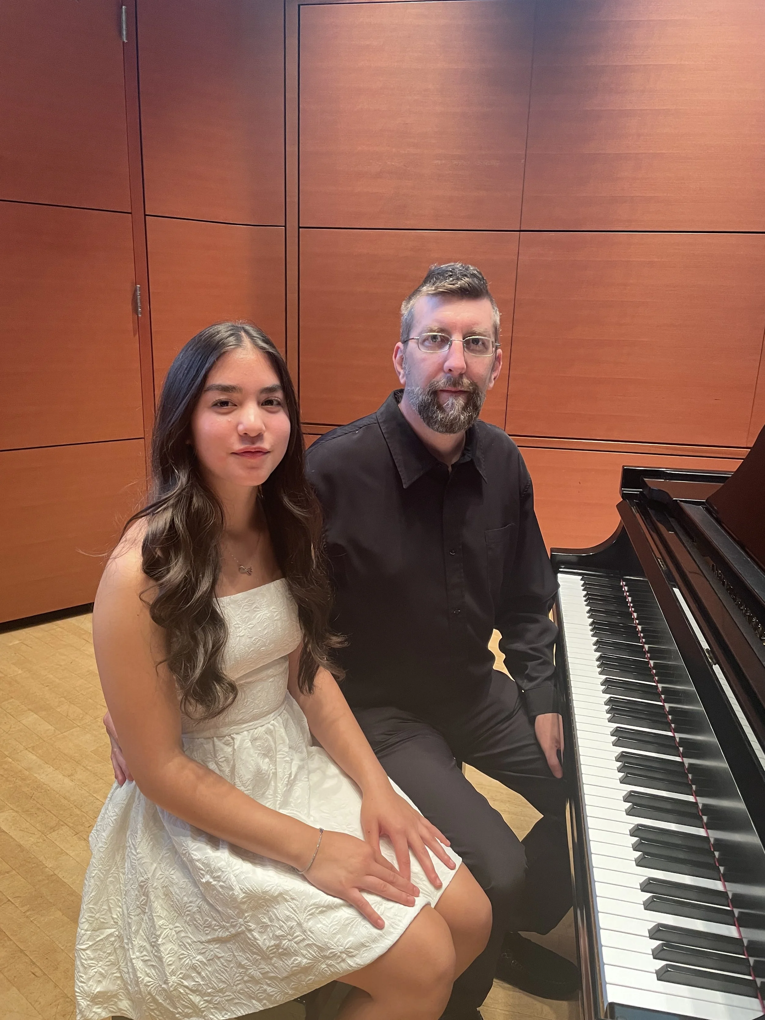 A young woman in a white dress sitting at a black grand piano next to a man in a black shirt, all in a wooden classroom or concert hall.