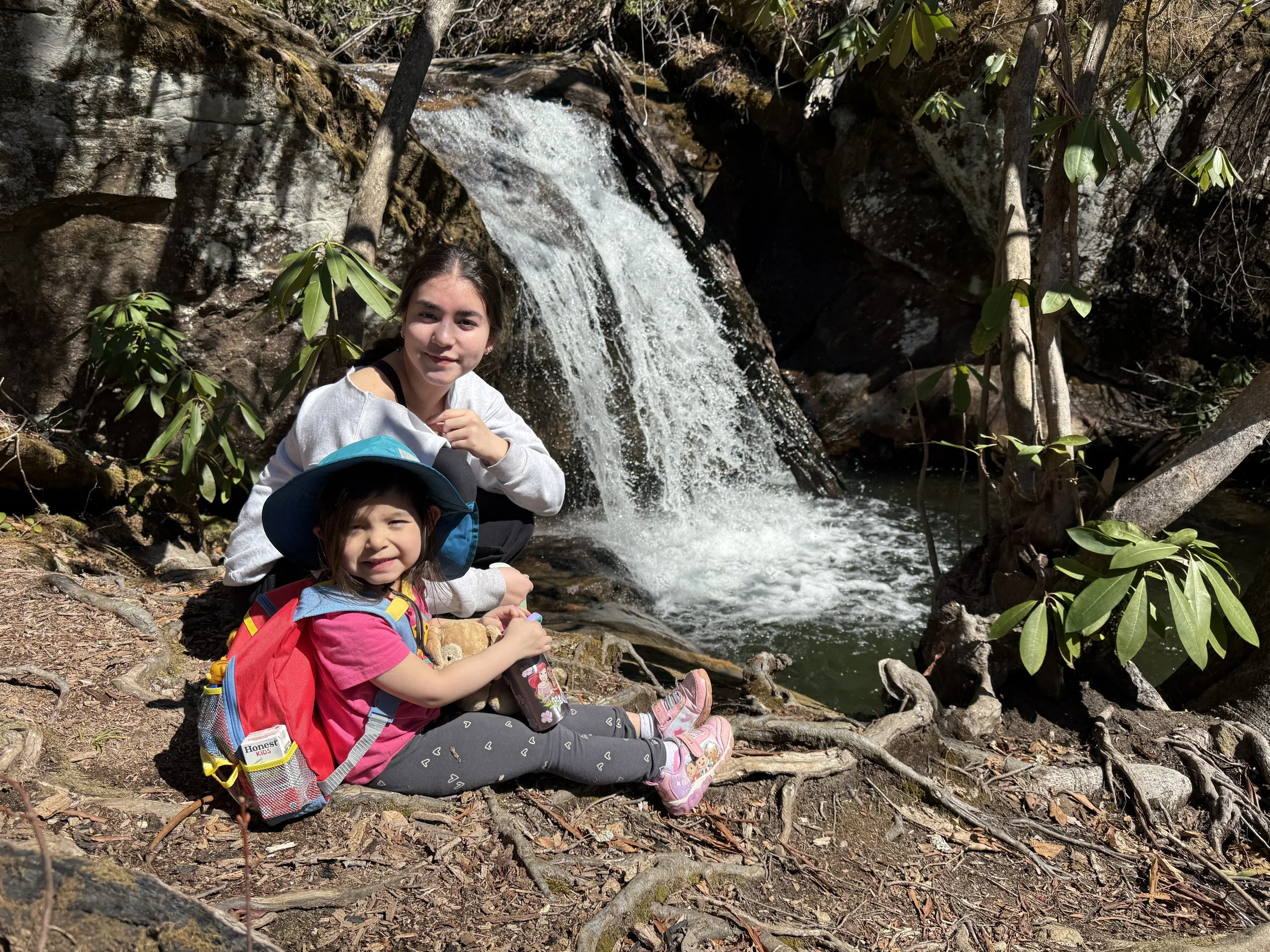 Two young girls sitting on the ground next to a small waterfall in a forest, with the older girl behind the younger. The younger girl wears a pink shirt, grey leggings with white hearts, pink shoes, a blue hat, and a red backpack. The older girl wear