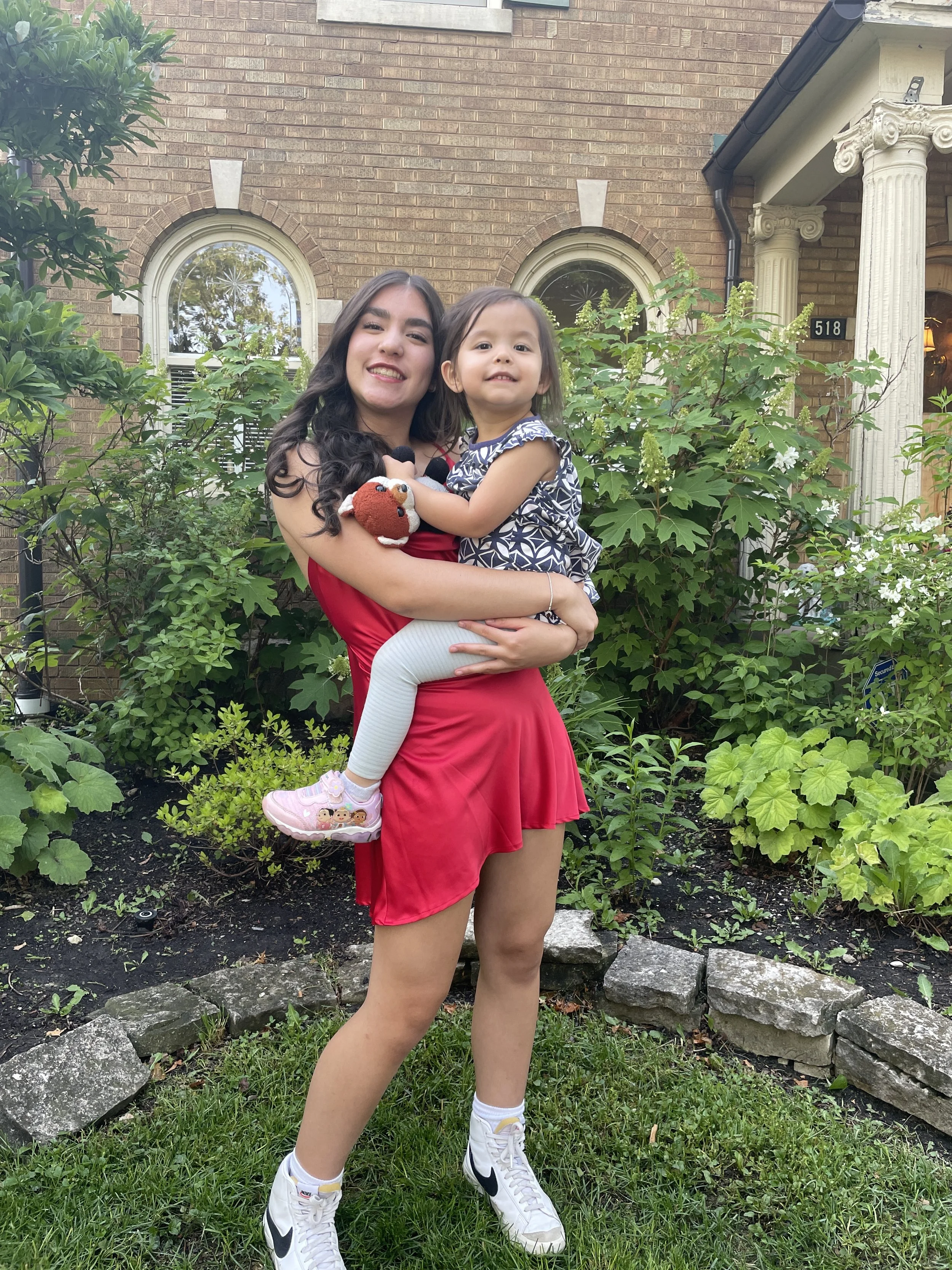 A young woman in a red dress holding a small girl in a patterned dress standing in front of a house with greenery and a brick facade.
