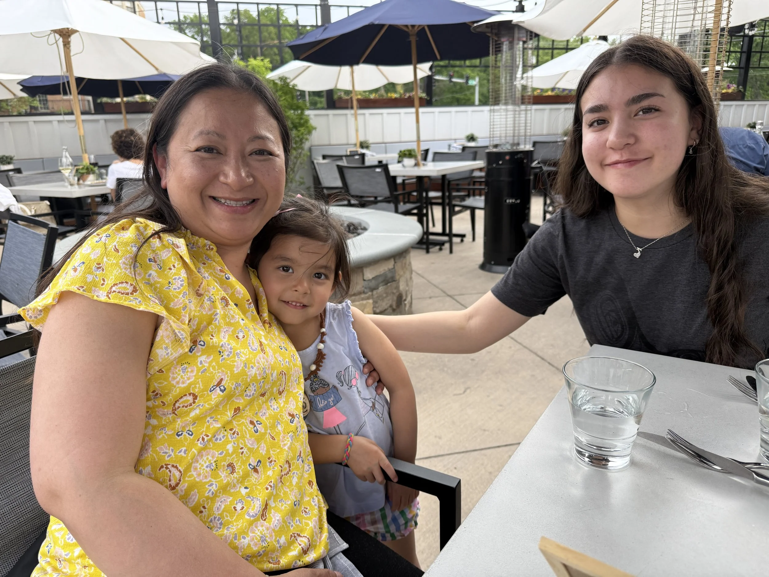 Three women, a middle-aged woman in a yellow floral top, a young girl in a white shirt, and a teenage girl in a dark gray shirt, are sitting at an outdoor restaurant table. The middle-aged woman and the young girl are smiling at the camera, with the