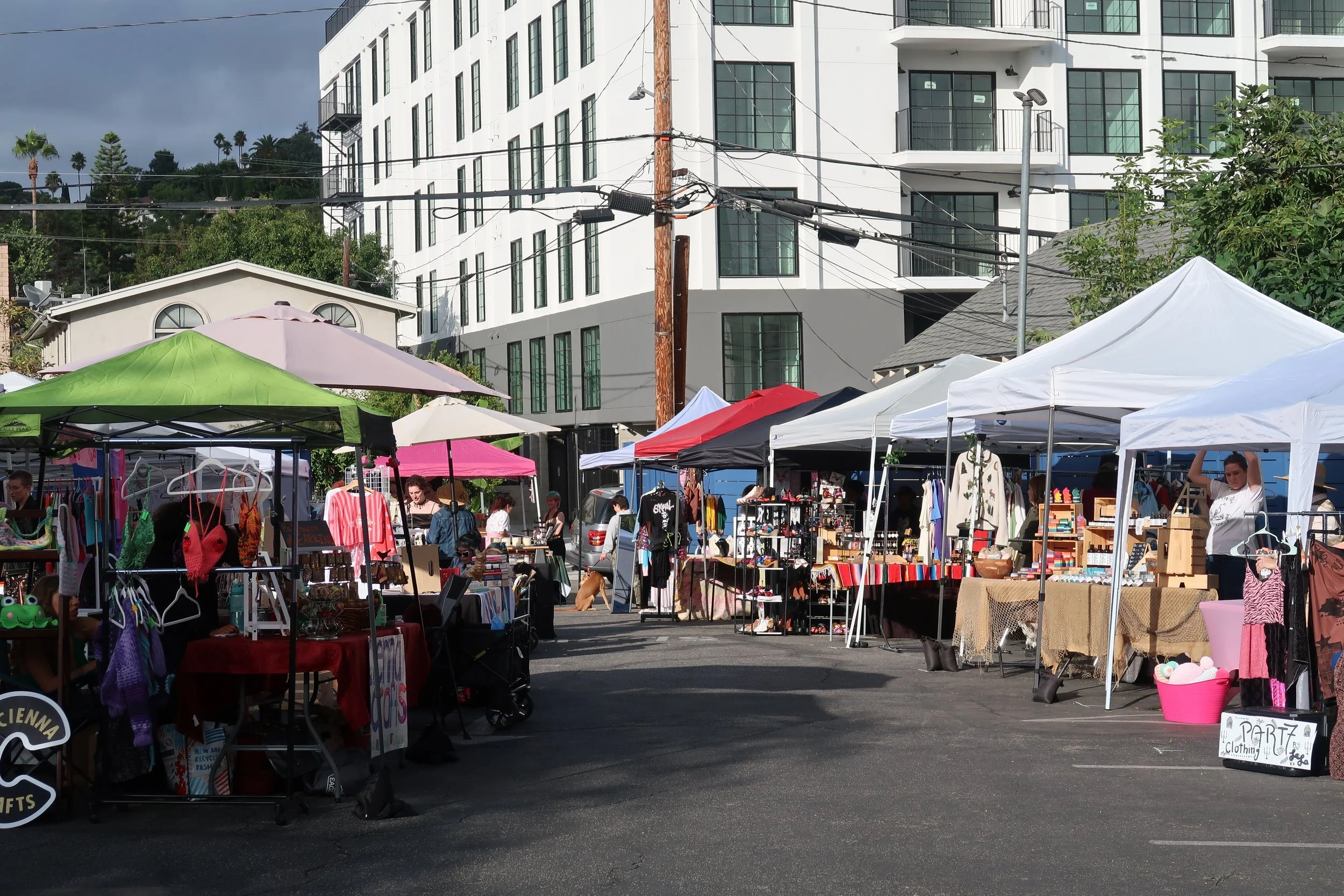 Outdoor market with vendor tents selling clothes, accessories, and crafts in a parking lot with modern building and utility poles in the background.