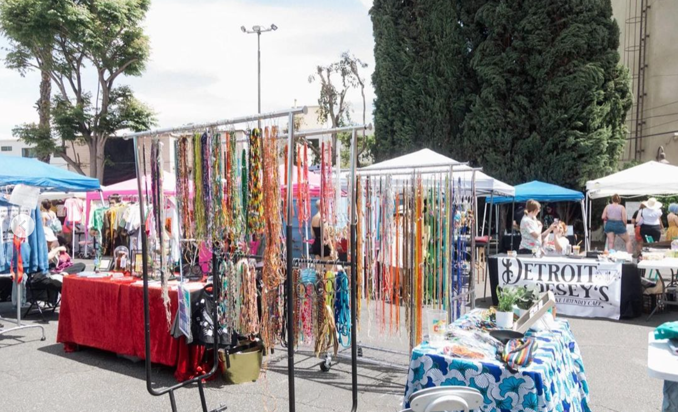 At a bustling outdoor flea market, there are various vendor tables with clothes, jewelry, and crafts. One table is draped with a red cloth displaying necklaces, while another table features rainbow-colored accessories. Tents provide shade, and people