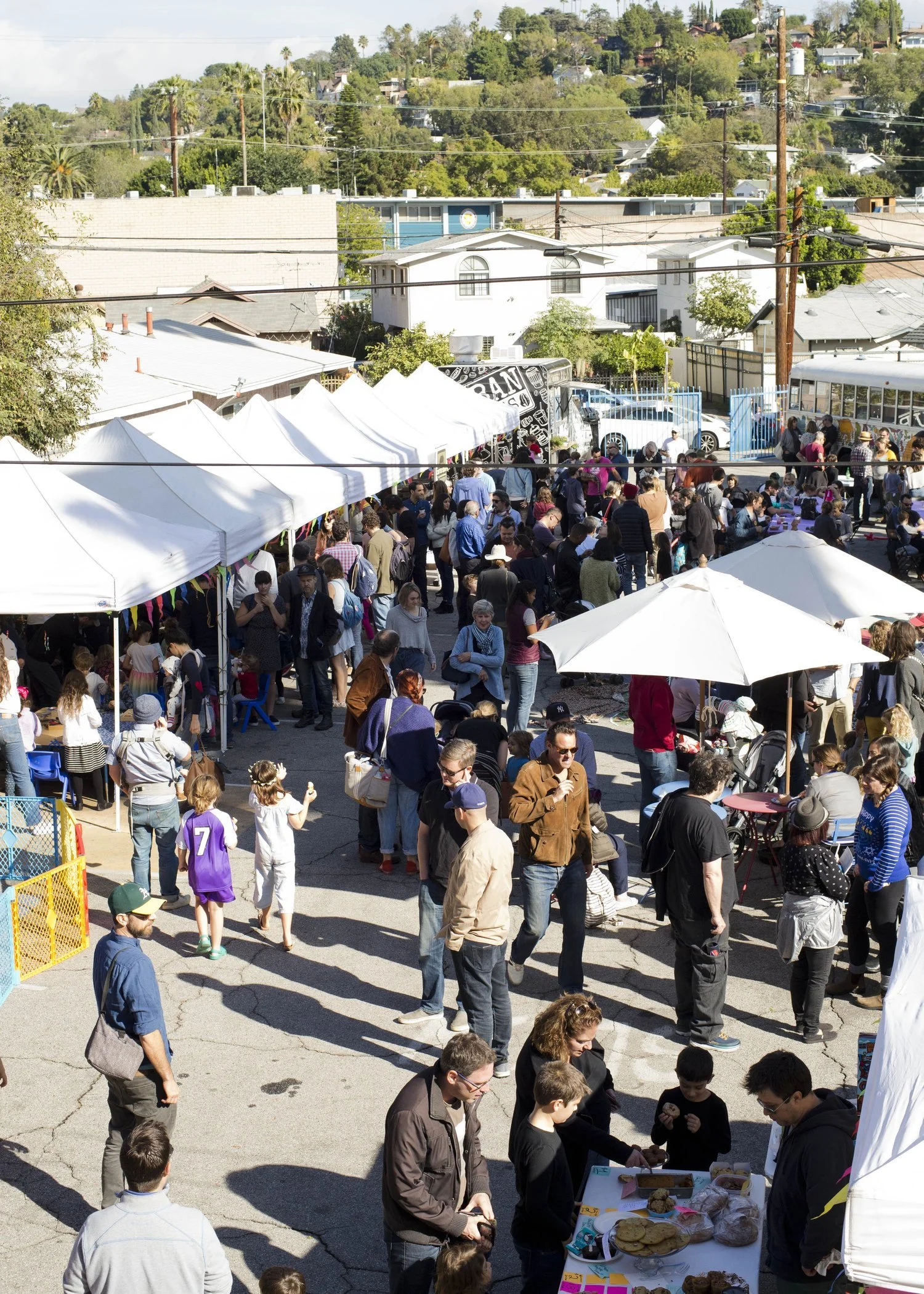Crowd of people at an outdoor market with white tents, tables, and umbrellas on a sunny day, surrounded by residential houses and green hills.