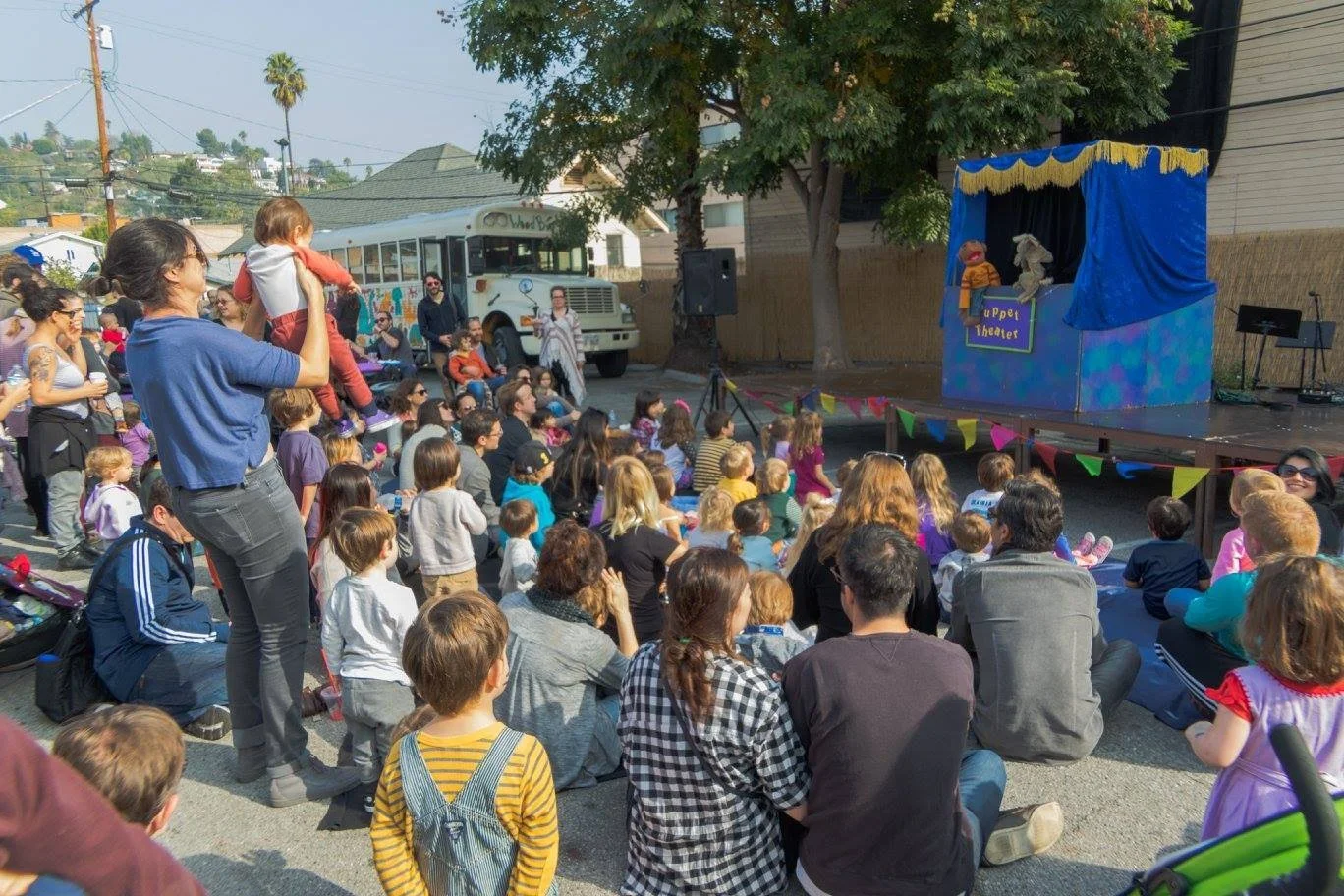 A puppet show is taking place outdoors with a large crowd of children and adults seated on the street, watching a stage with puppets and a sign that reads "Puppet Theater."