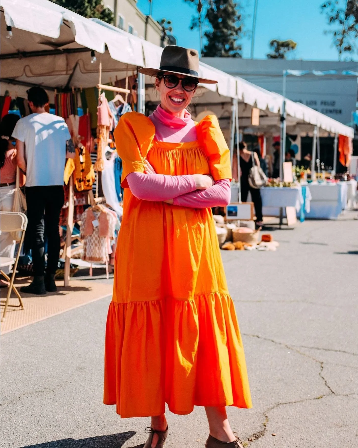 Woman in an orange dress and pink long-sleeve shirt, wearing a wide-brimmed hat and sunglasses, smiling at an outdoor market.