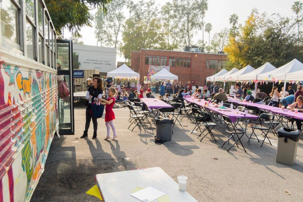 An outdoor community event with a colorful food truck, several white tents, and people sitting at tables decorated with pink tablecloths, set up in a parking lot with a building labeled 'Hollywood Los Feliz Jewish Community Center' in the background.
