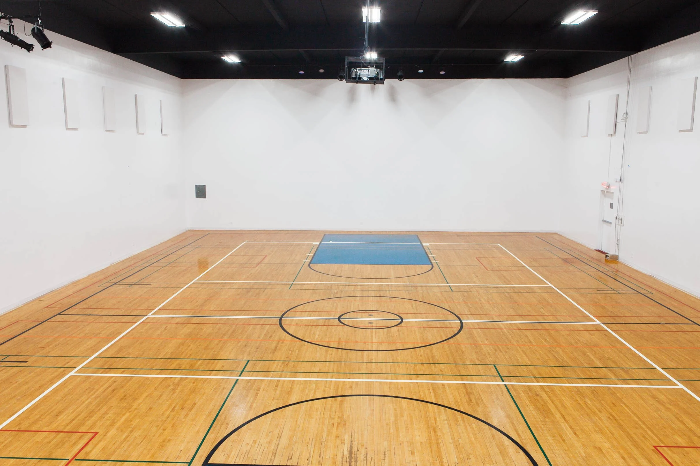 Empty indoor basketball court with wooden flooring, white walls, and black ceiling with lights.