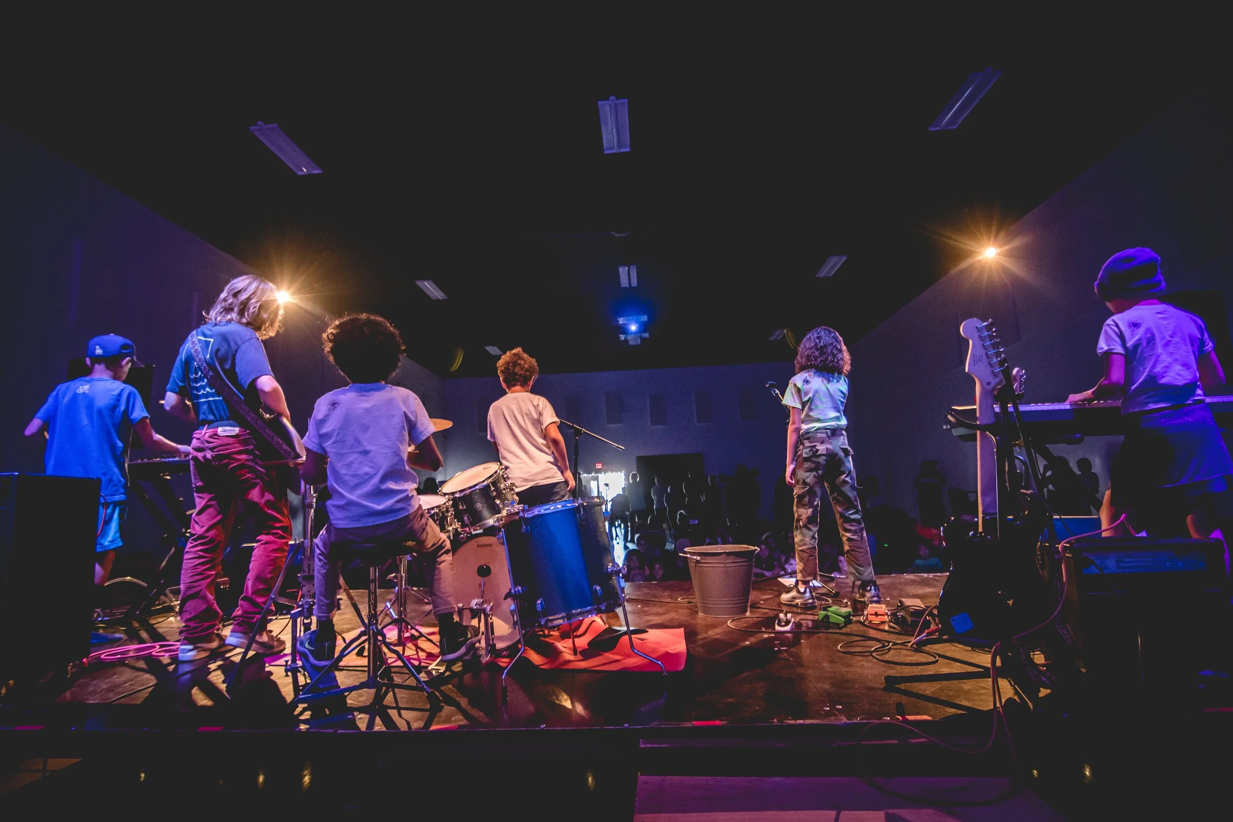 Children performing on stage with musical instruments, in a dark room with stage lighting.