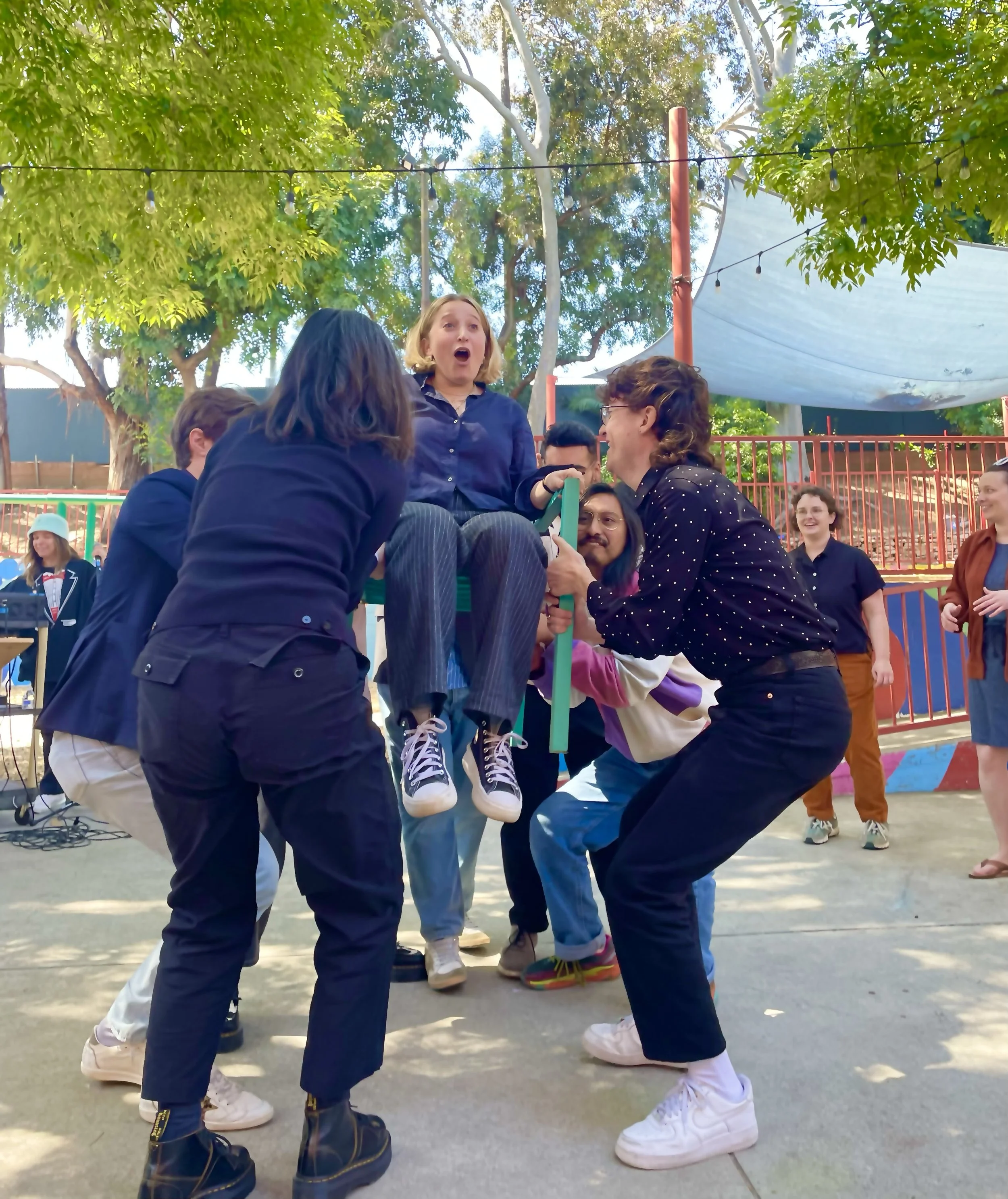 People celebrating outdoors, lifting a young woman in a chair in the air, with joyful expressions, trees, and a canopy overhead.