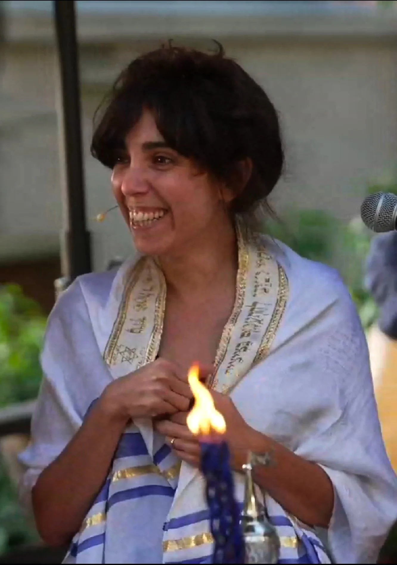 A woman with short dark hair smiling and holding her hands over her chest. She is dressed in white with gold and blue accents, and there is a lit candle in the foreground.