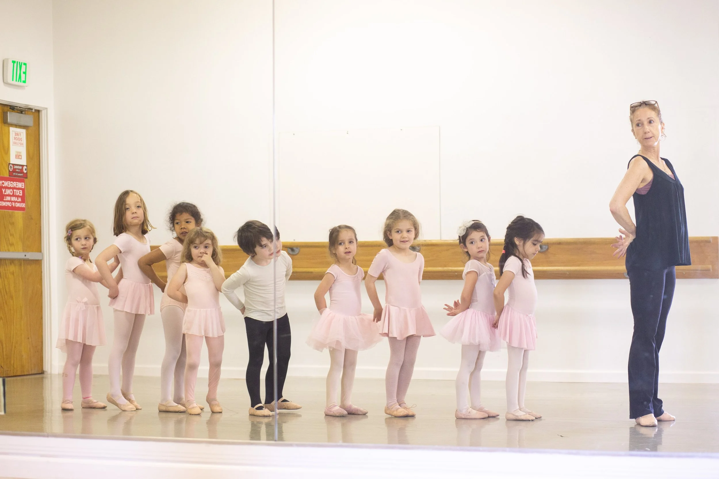 A dance teacher instructing a group of young girls in pink ballet outfits in a dance studio with a mirrored wall.