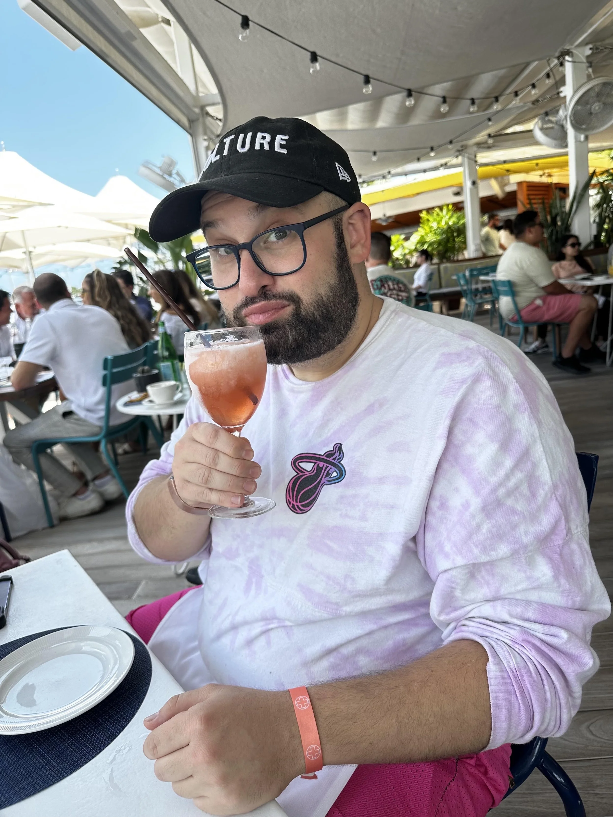 A man with glasses and a beard wearing a black cap that says 'CULTURE' and a tie-dye hoodie, sitting at an outdoor restaurant, holding a pink cocktail in a stemmed glass with a straw, surrounded by other diners under a shaded canopy.