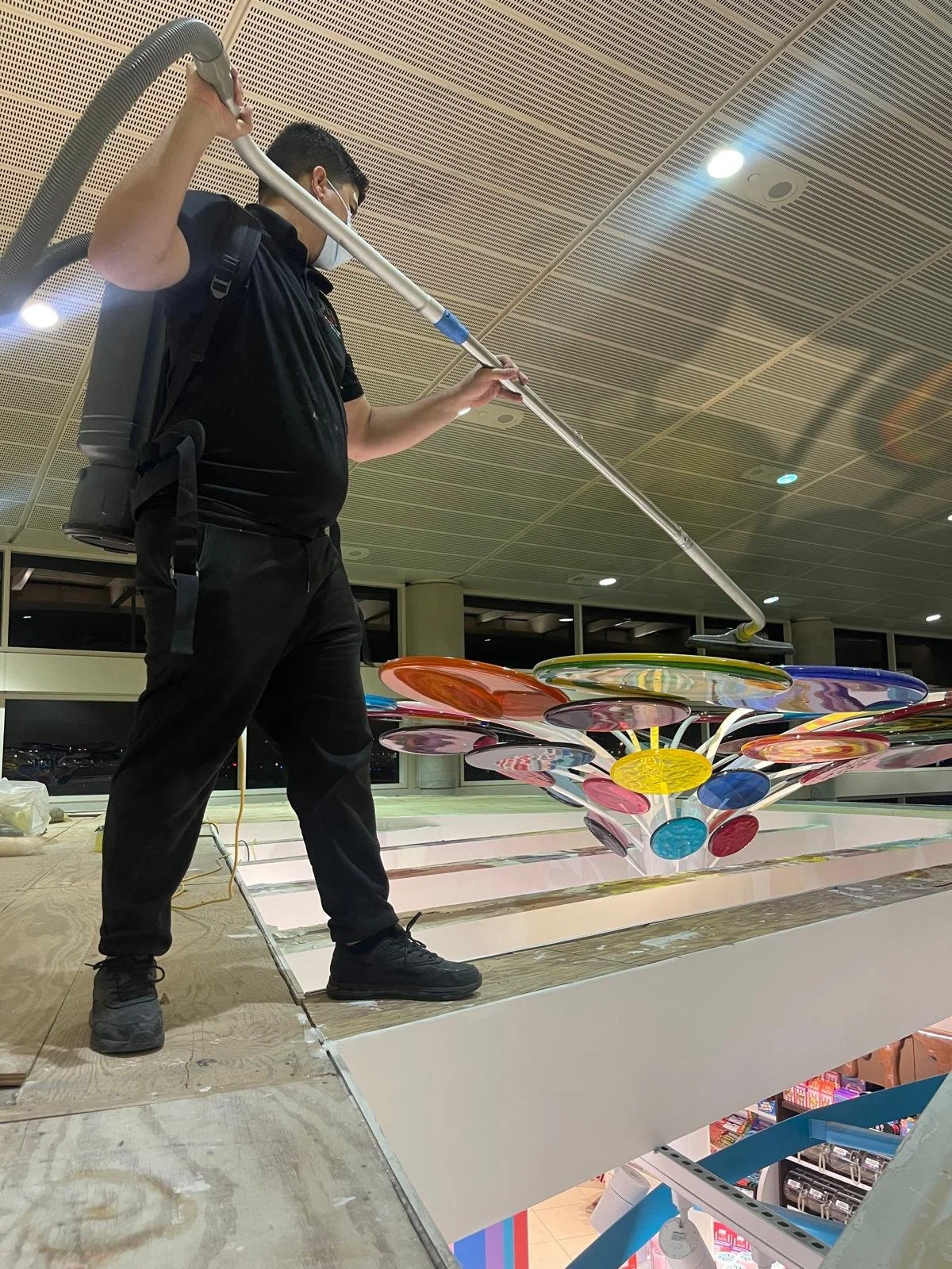 A man wearing glasses, a black shirt, and face mask cleaning a colorful, large decorative sculpture using a long vacuum cleaner at an indoor public space.