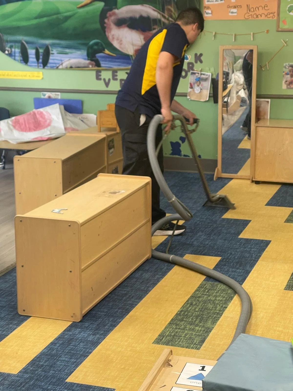 A man vacuuming a colorful carpet in a children's playroom or classroom. The room has wooden furniture, posters, and a mirror on the wall.