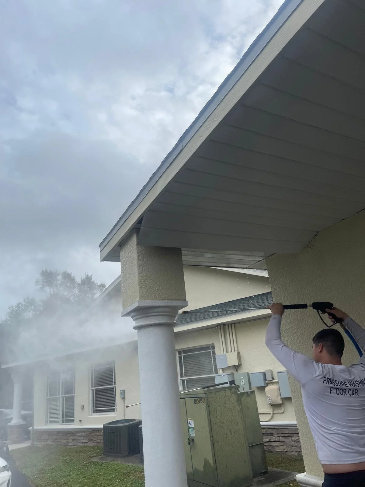 A man pressure washing the side of a house under the roof, with water spraying onto the exterior wall.