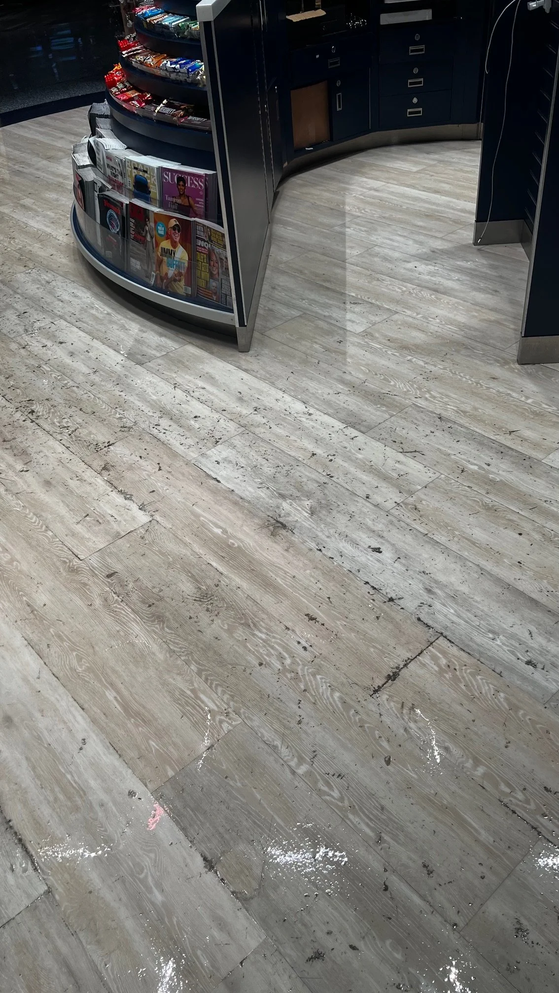 A magazine and snack display area next to a curved checkout counter in a store with light-colored wooden flooring, some chalk marks, and scattered debris.