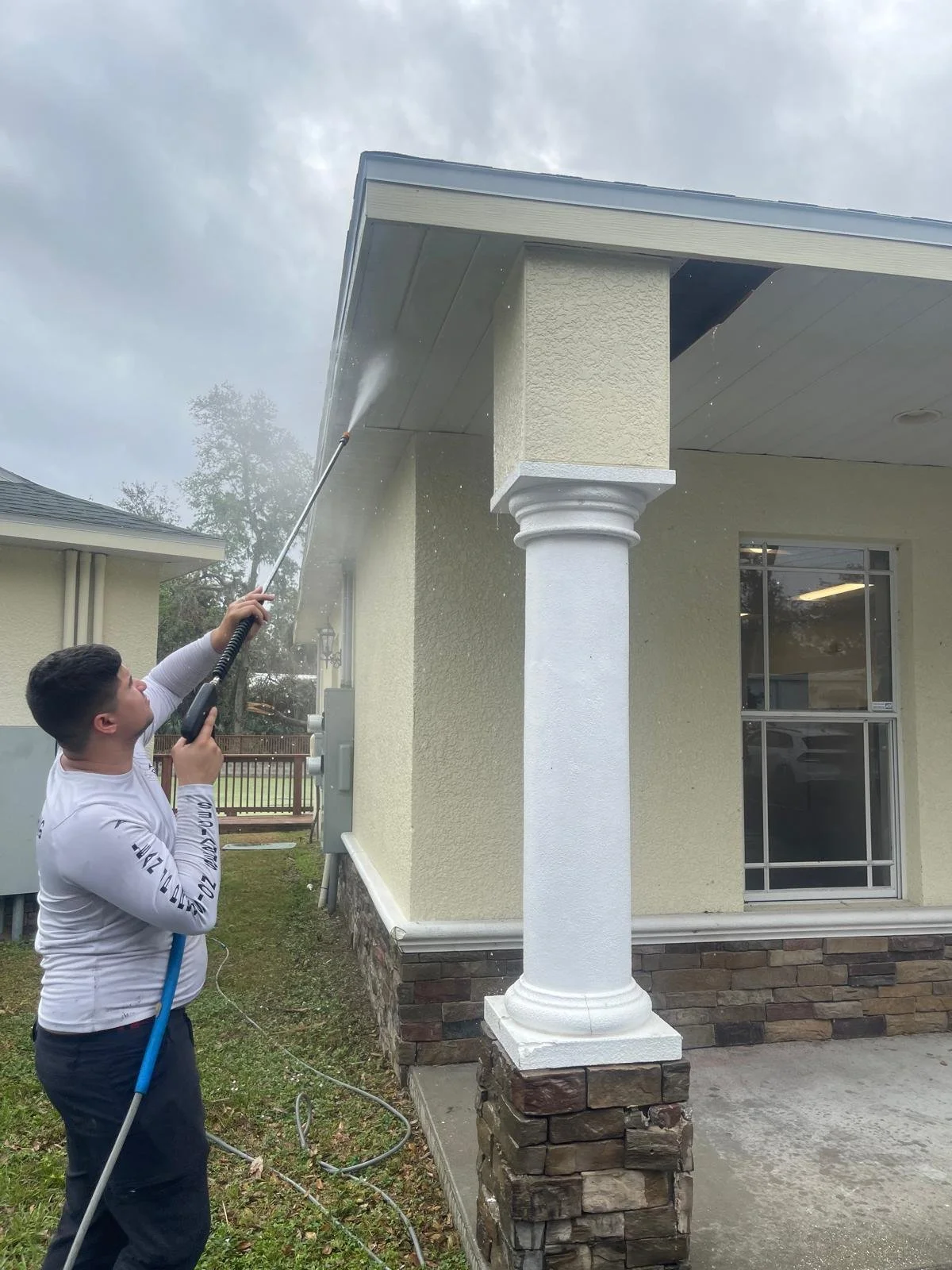 A man is pressure washing the exterior of a house, focusing on the roof overhang and a column supporting the porch.