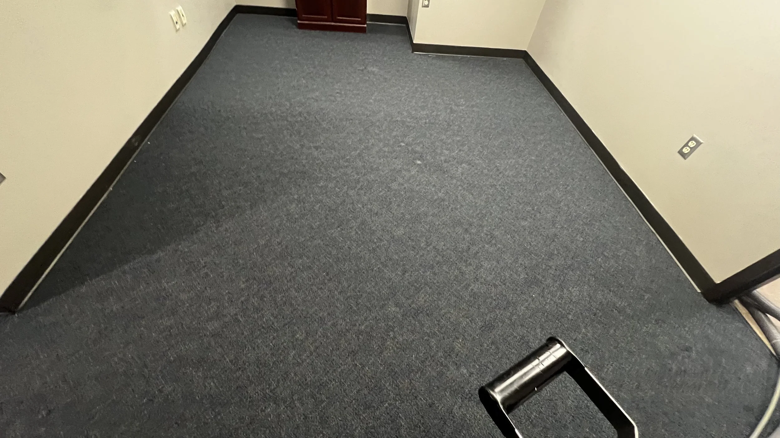 Empty office with gray carpet, beige walls, a wooden cabinet, black baseboards, and electrical outlets, with part of a black chair visible at the bottom right.