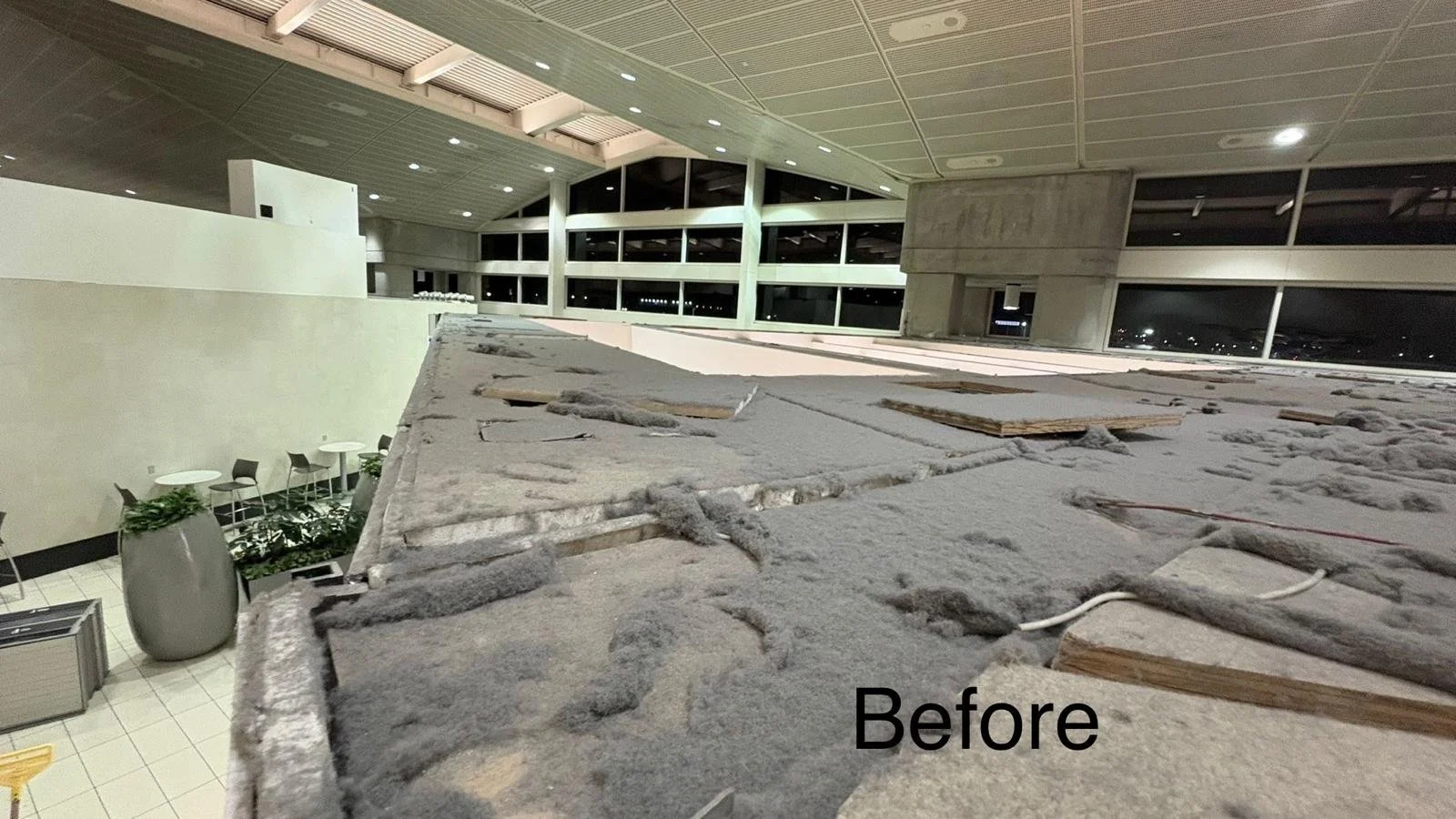 Interior of an airport terminal under renovation with unfinished flooring, visible insulation, and construction materials. The space is well-lit with large windows showing a night scene outside.