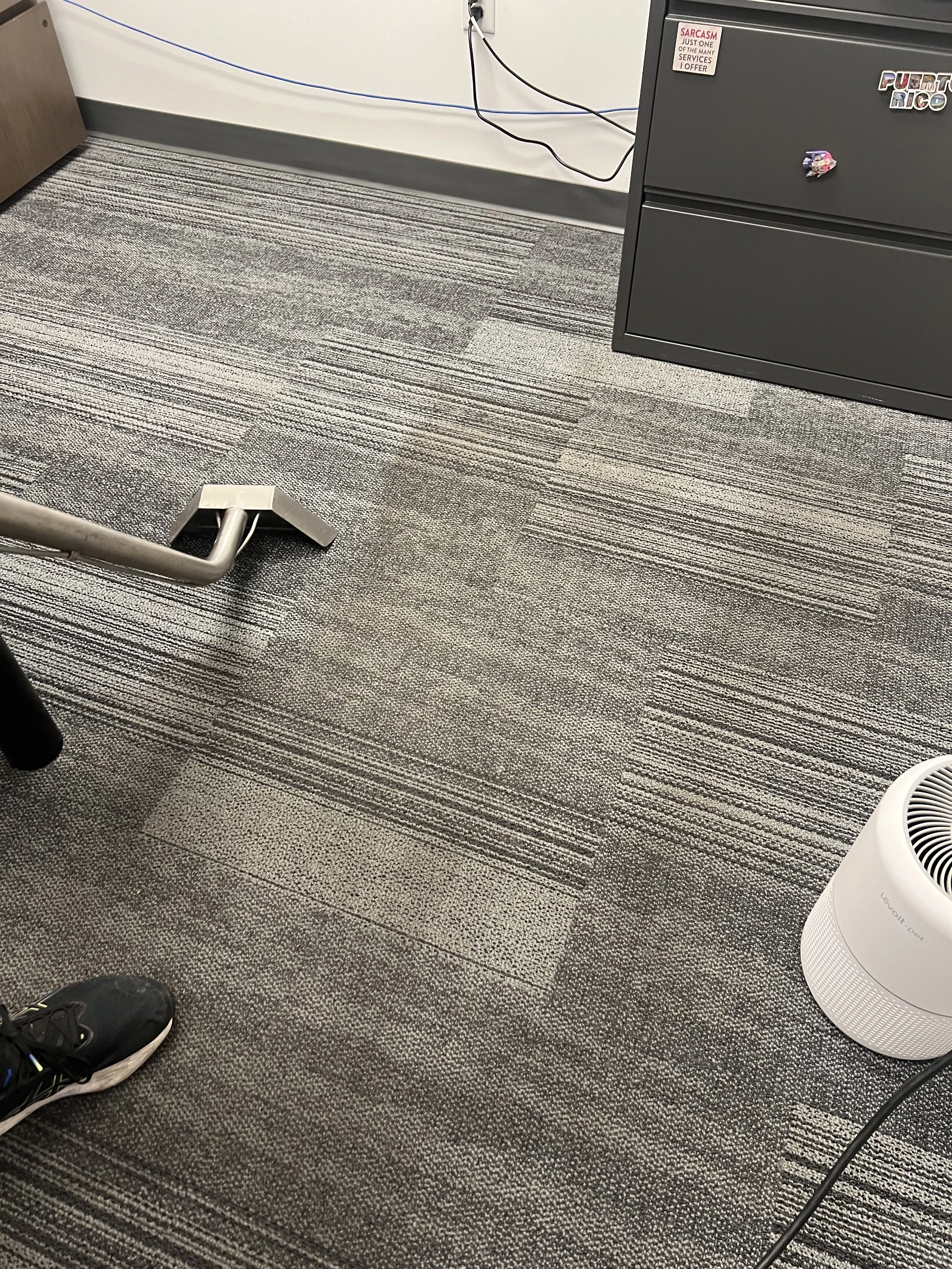 Office carpeted floor with a person's black athletic shoe, a metal chair leg, a white indoor air purifier, a grey filing cabinet with stickers, and visible electrical cords.