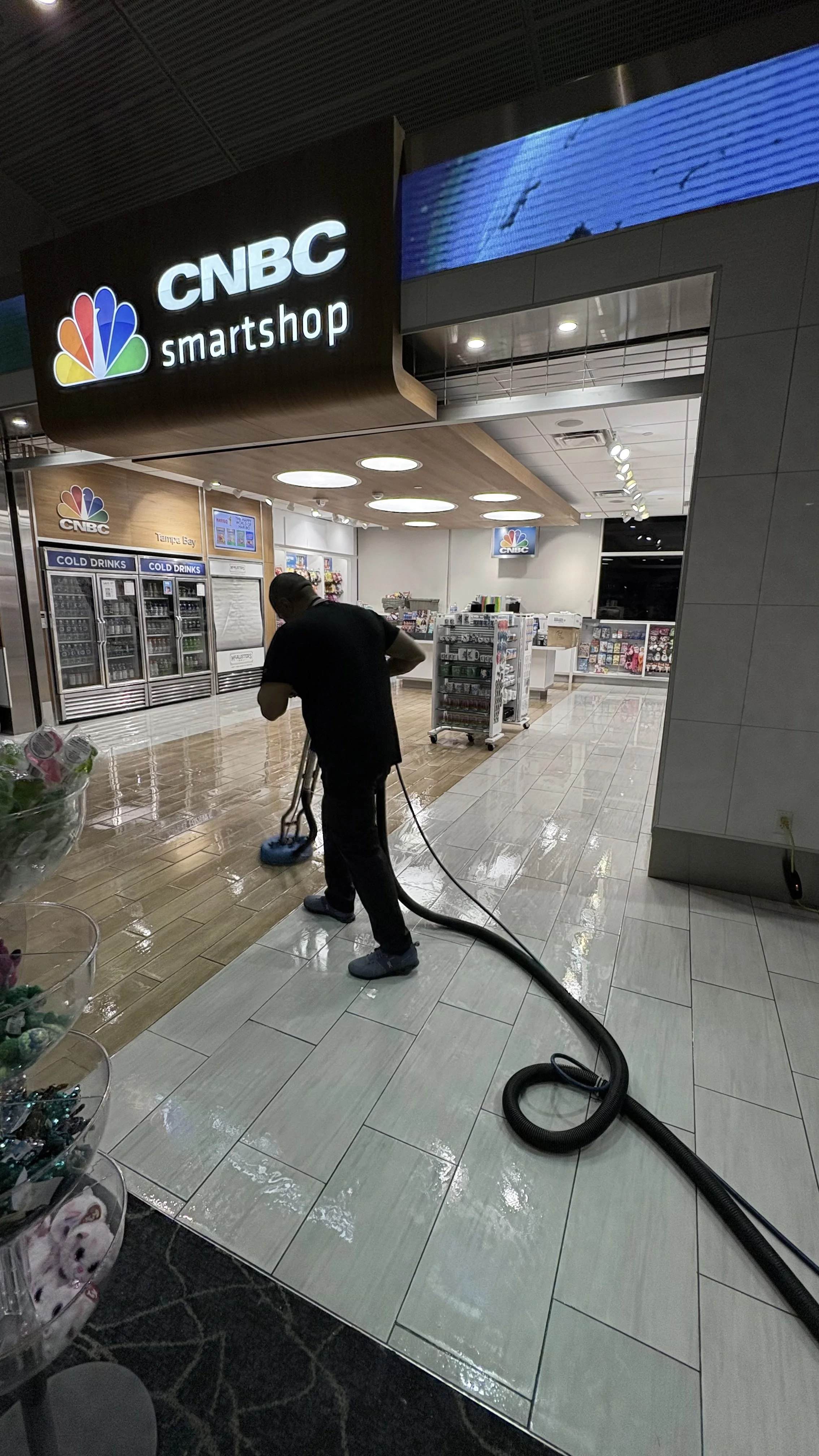 A person cleaning or scrubbing the tiled floor of a CNBC SmartShop store with a vacuum or cleaning machine, in a brightly lit indoor shopping area.