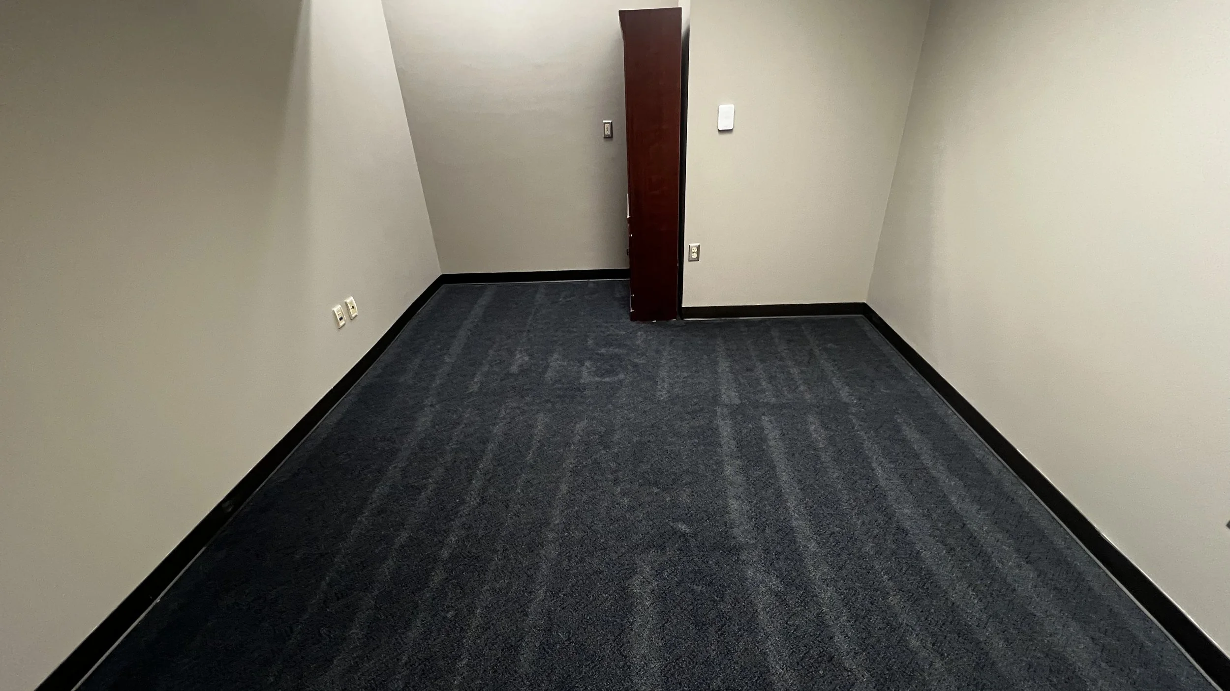 Empty office room with beige walls, dark gray carpet, black baseboards, and a wooden door.