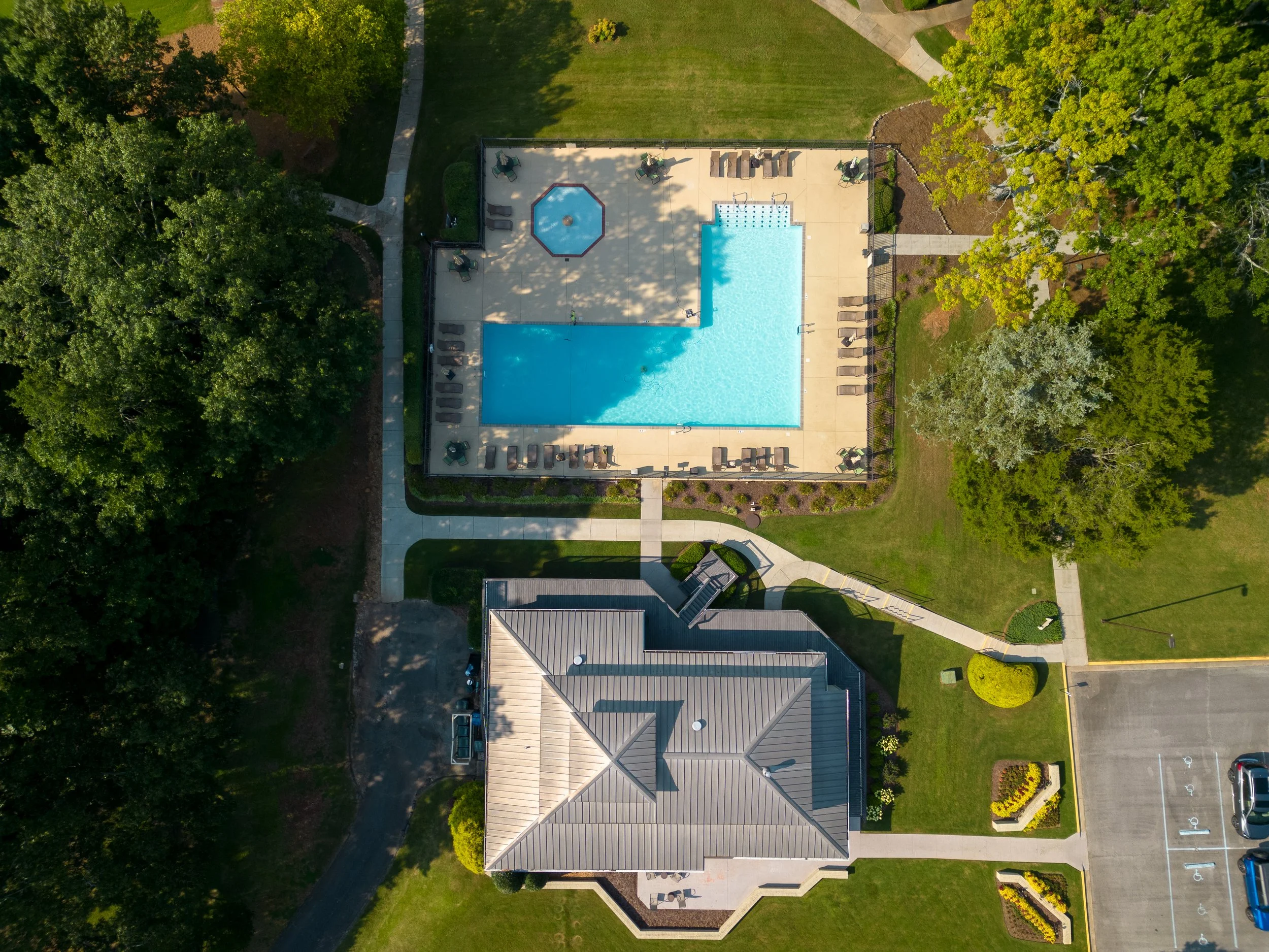 Aerial view of a swimming pool area with surrounding patio chairs, a small round hot tub, walking paths, green trees, and a building with a gray roof, adjacent to a parking lot with marked parking spaces.