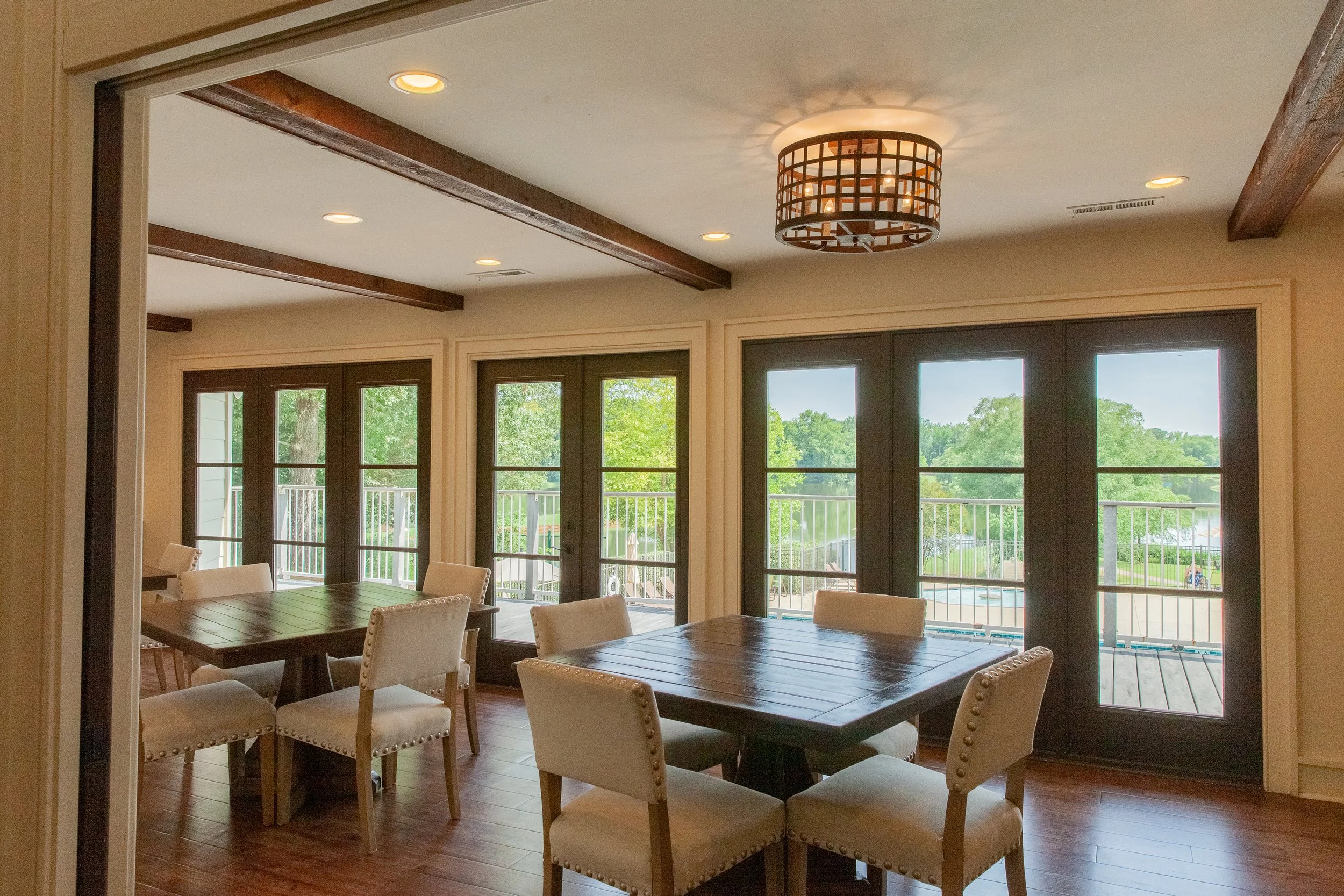 Dining room with wooden tables, upholstered chairs, large windows, and a view of a backyard with a patio and greenery.