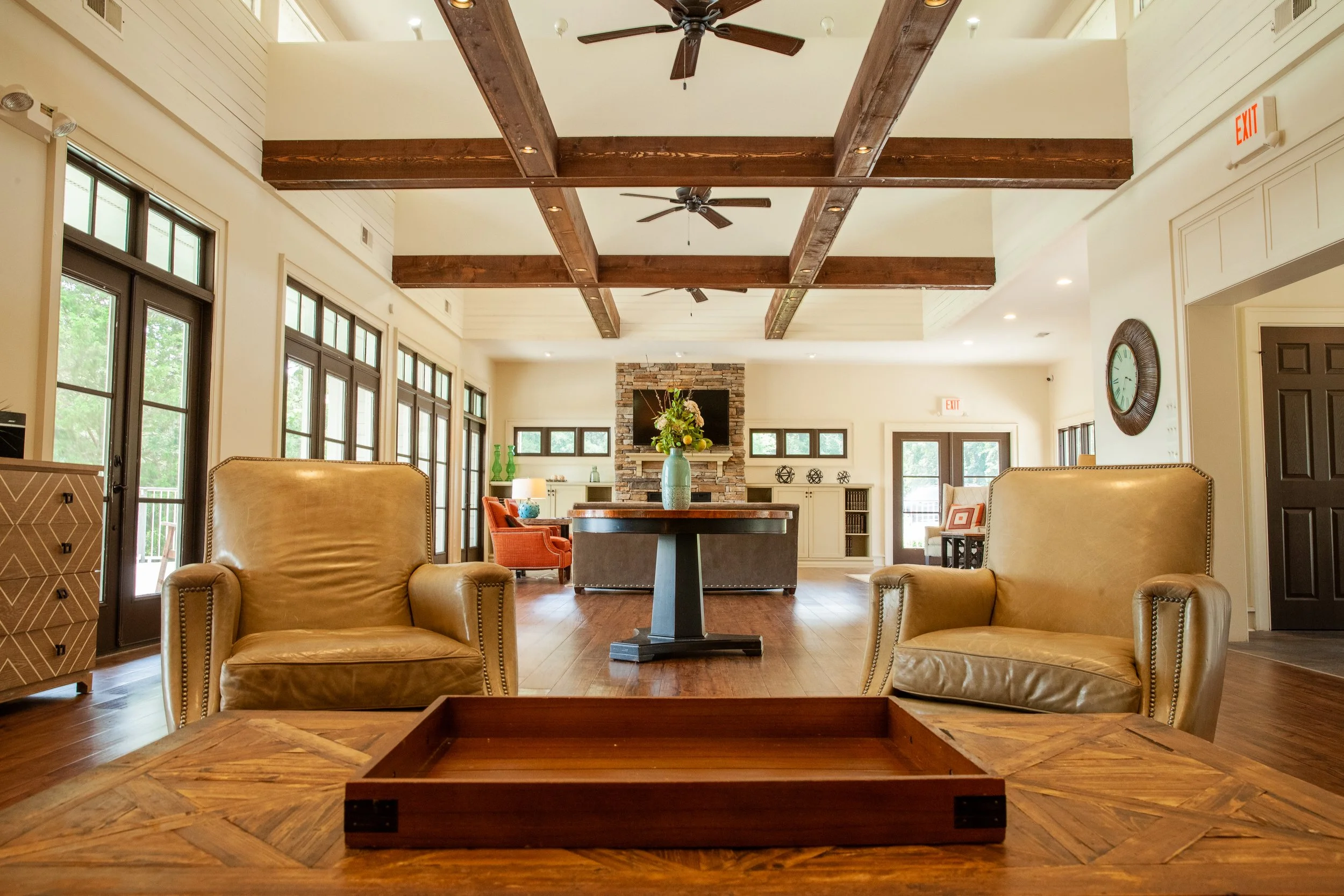 Spacious living room with beige leather armchairs, a wooden table with a tray, large windows, and a stone fireplace with a TV above, decorated with a large flower arrangement in the center.