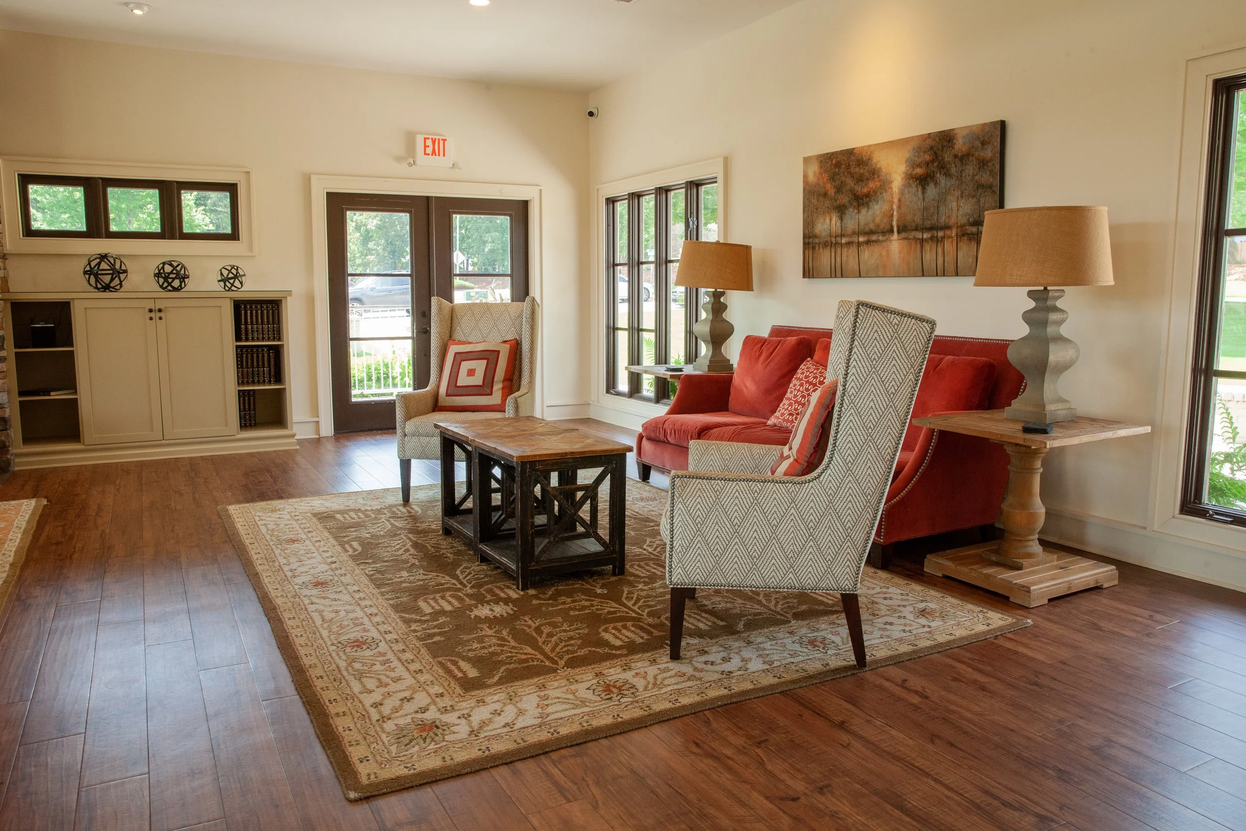 Living room with hardwood floors, a red sofa, two patterned armchairs, a wooden coffee table, and two lamps on side tables. There is a large painting on the wall, and windows letting in natural light.