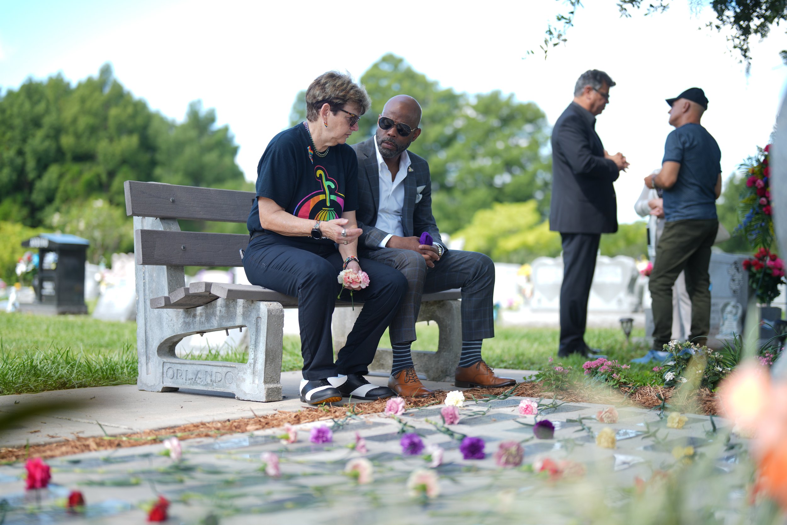 People paying respects at a gravesite with rose petals and flowers on the ground, people standing and talking nearby, trees in the background.