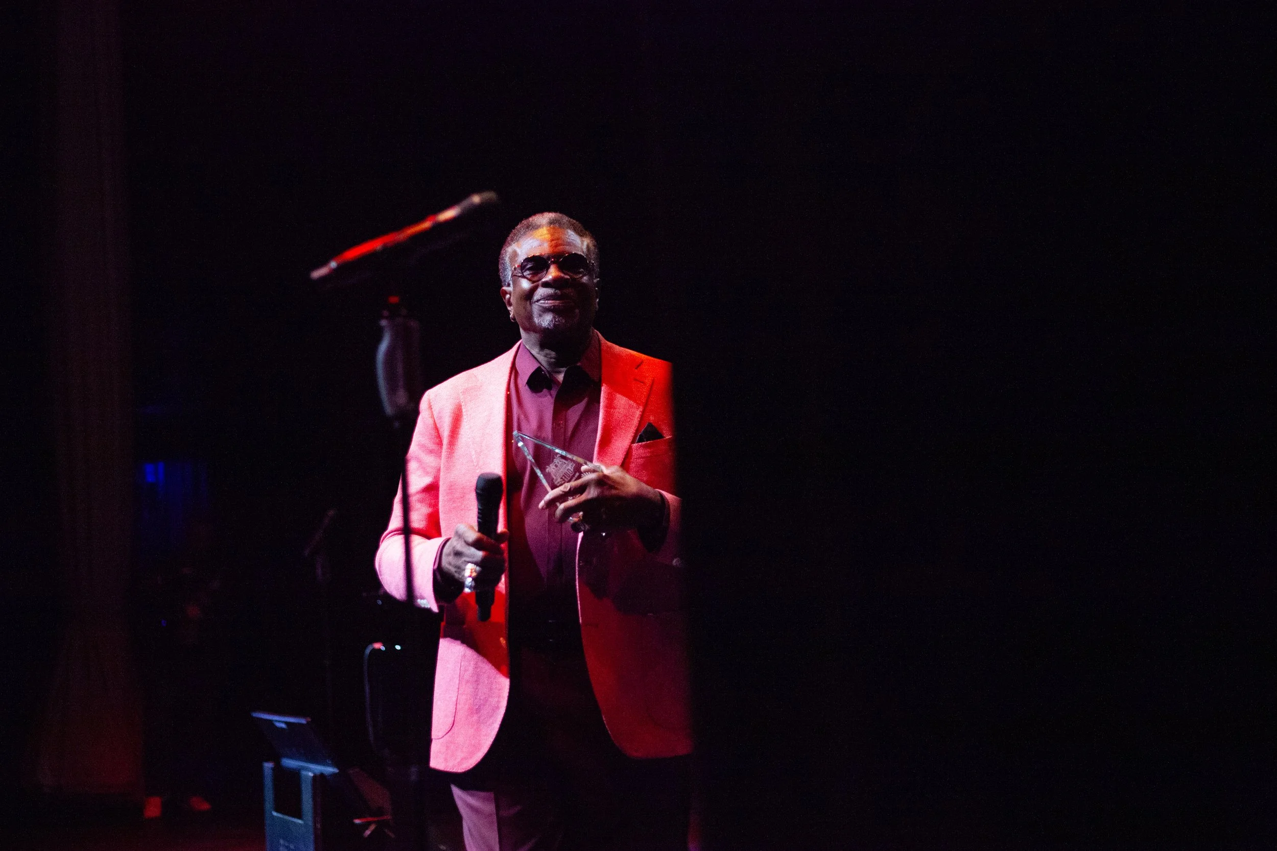 A man in a red suit and sunglasses holding a microphone and a glass award on a dark stage.