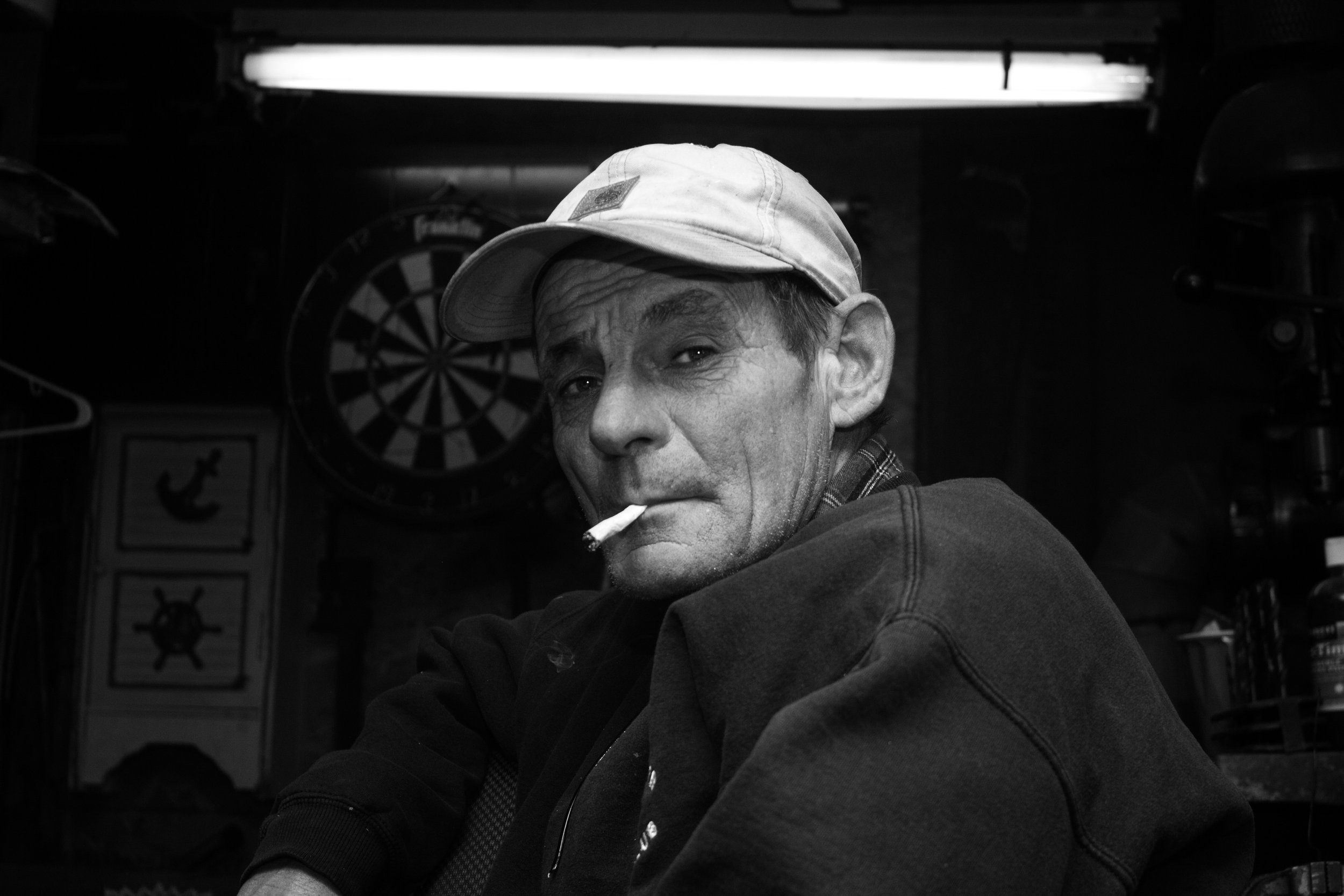 A black-and-white photo of a man with a cigarette in his mouth, wearing a cap and a dark sweatshirt, sitting in a dimly lit room with a dartboard and nautical-themed decor in the background.