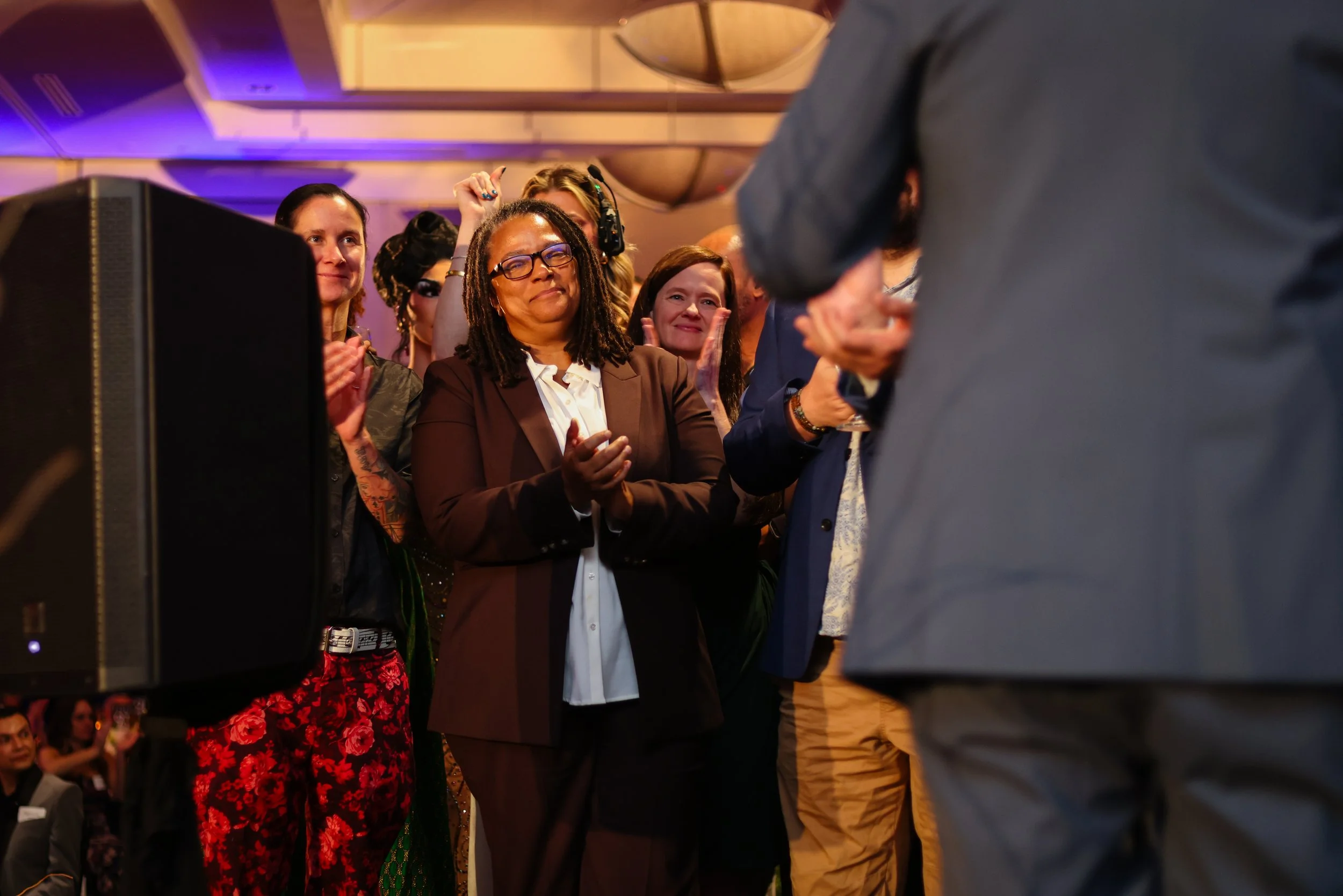 Group of people at an event, including a woman with glasses and dreadlocks in a brown suit clapping, surrounded by others who are also clapping and smiling, in a well-lit indoor setting.