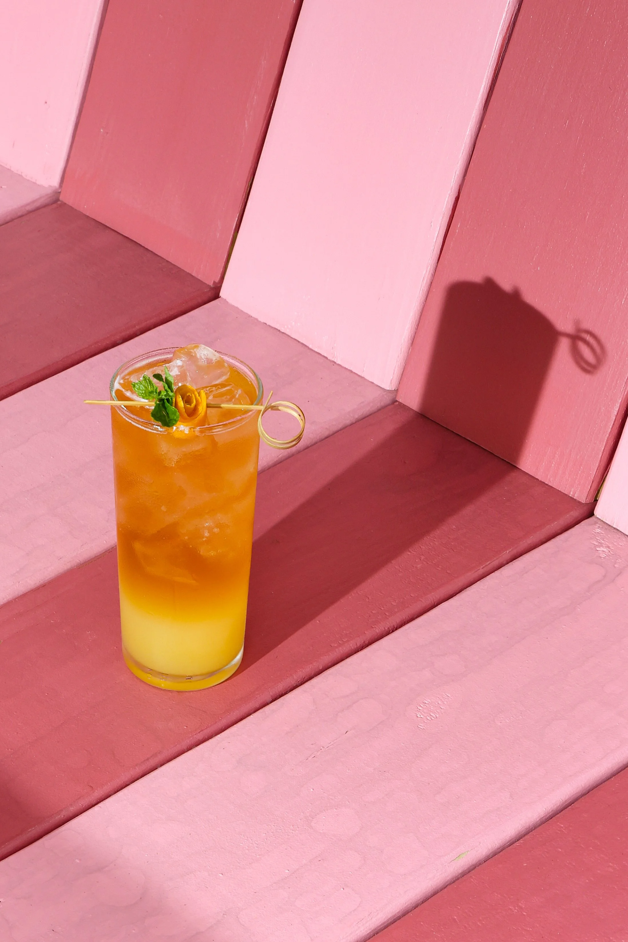 A tall yellow-orange layered cocktail in a clear glass with ice, garnished with a small yellow flower, mint leaf, and a decorative yellow straw, sitting on a pink wooden bench.