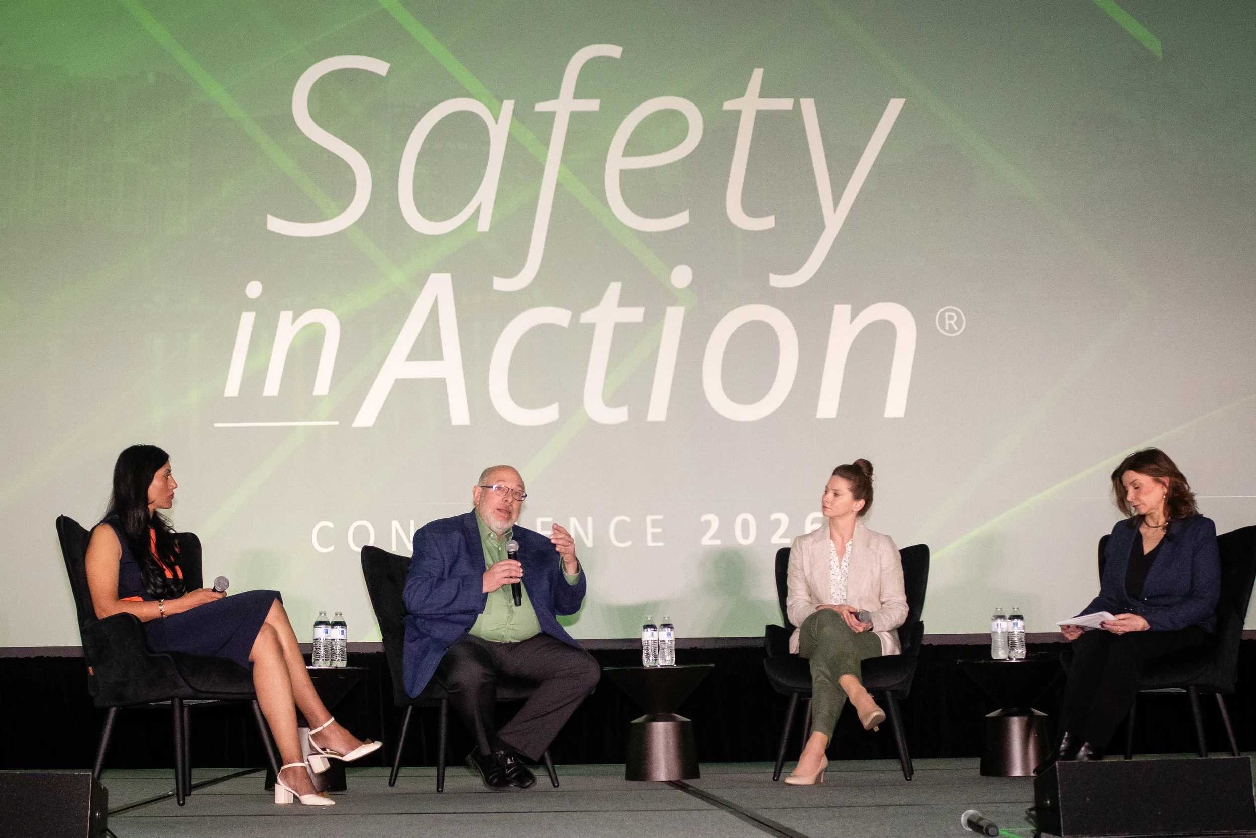 Panel discussion at a conference with four people seated on stage. A large screen behind them displays the words 'Safety in Action Conference 2023'. The panelists include three women and one man, each holding a microphone, with water bottles in front