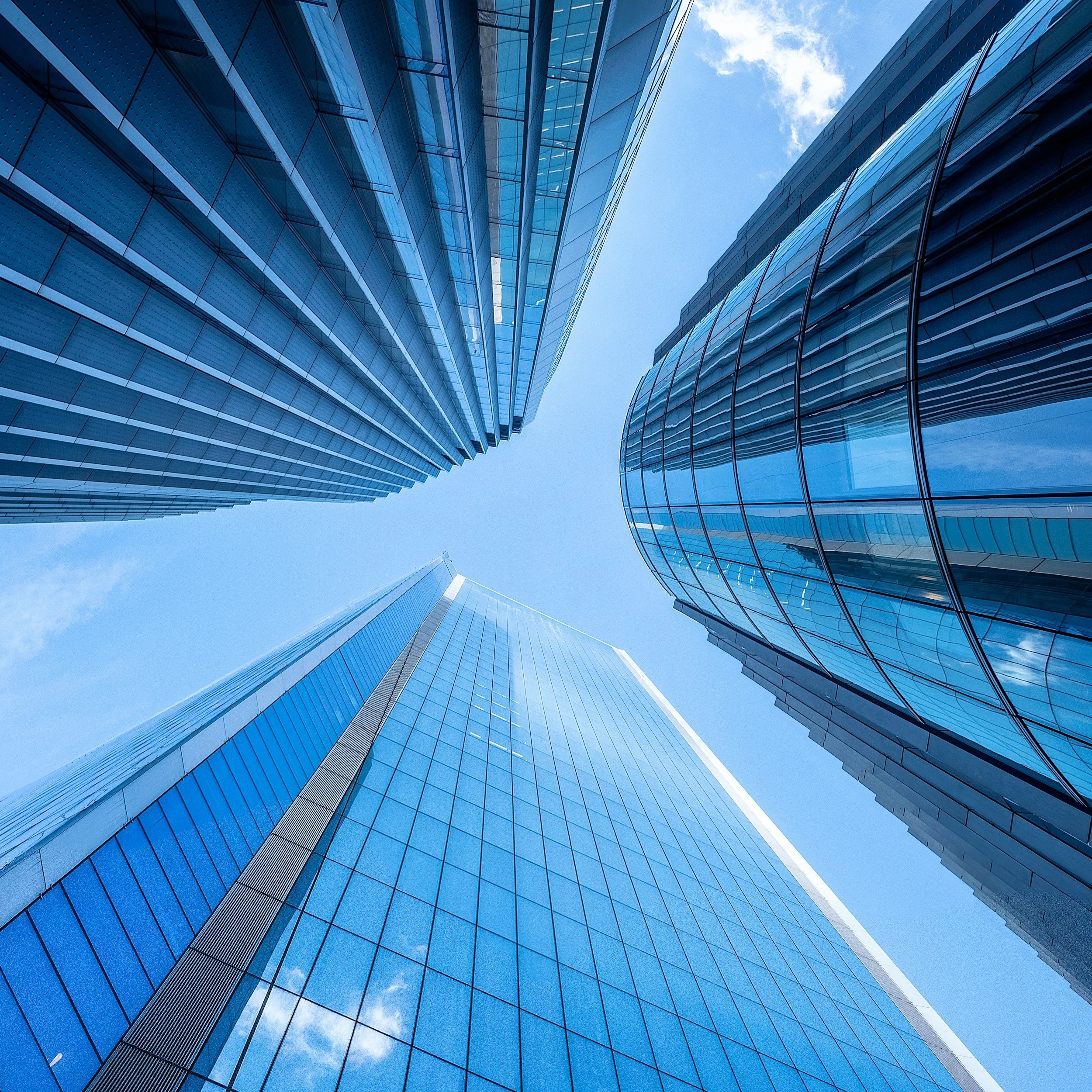 Looking up at the tall glass skyscrapers in a city, showing reflections of the sky and clouds on their blue-tinted glass facades.