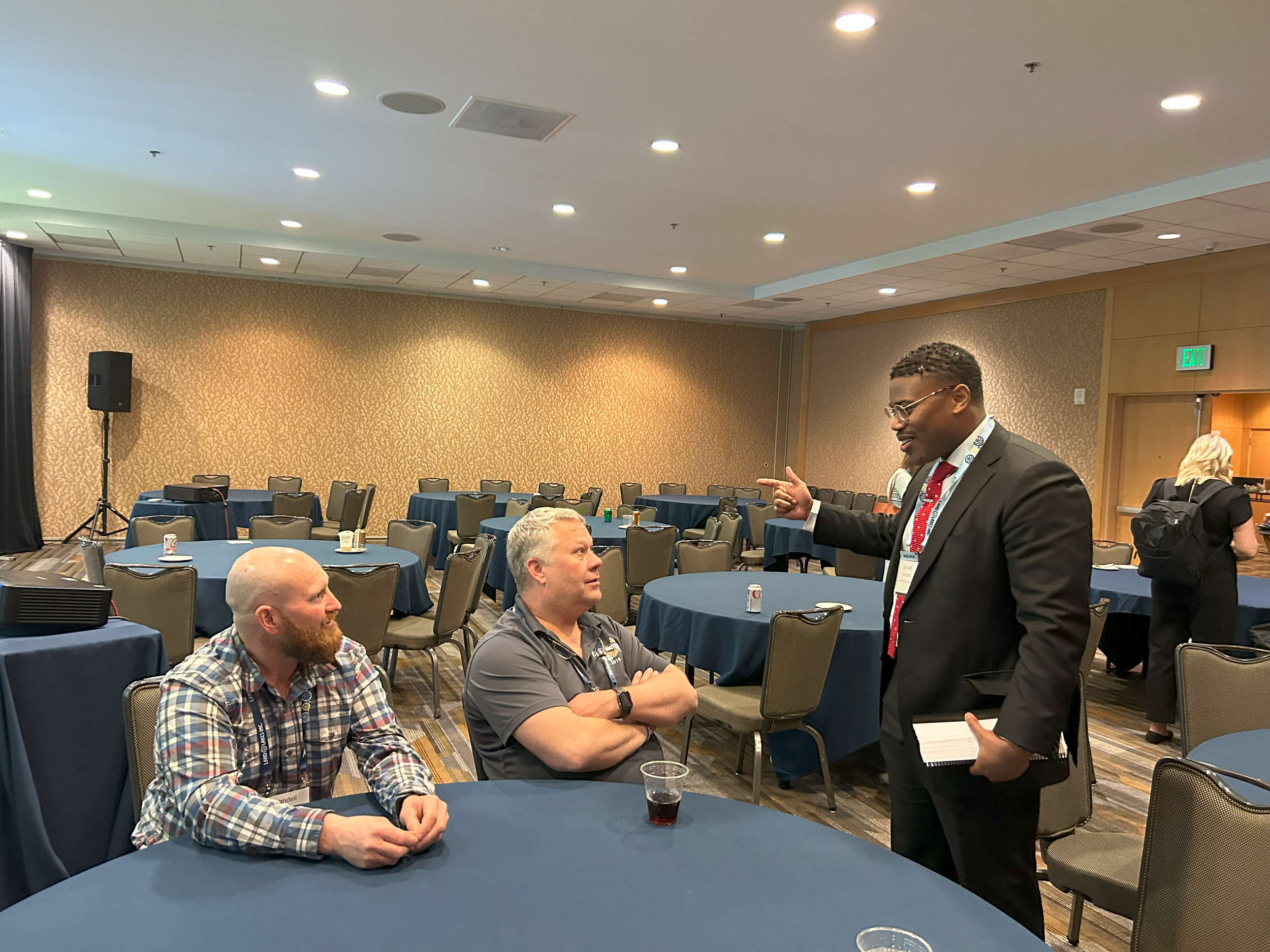 Three men engaged in conversation in a conference room. One man standing and pointing, two men sitting and listening. The room has round tables with blue tablecloths and empty chairs, with a sound system and exit sign visible.