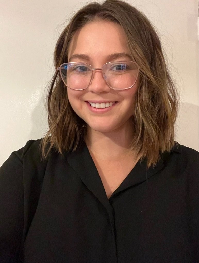 A young woman with shoulder-length brown hair, wearing glasses and a black shirt, smiling at the camera against a plain light-colored background.