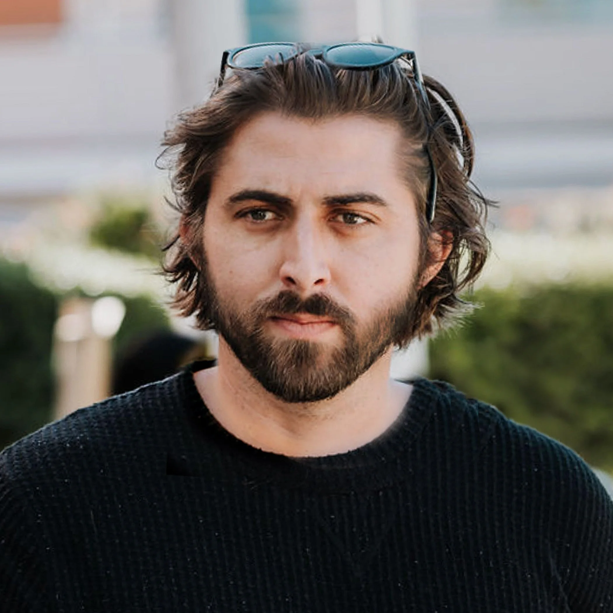 A man with medium-length wavy hair, a beard, and sunglasses on his head, wearing a black sweater outdoors.