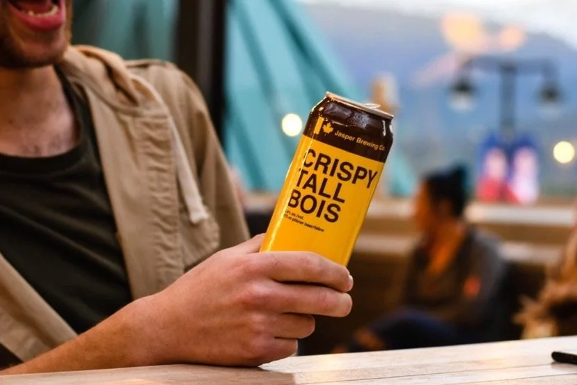 Person holding a can of Crispy Tall Bois beer at a wooden table in a bar or restaurant.