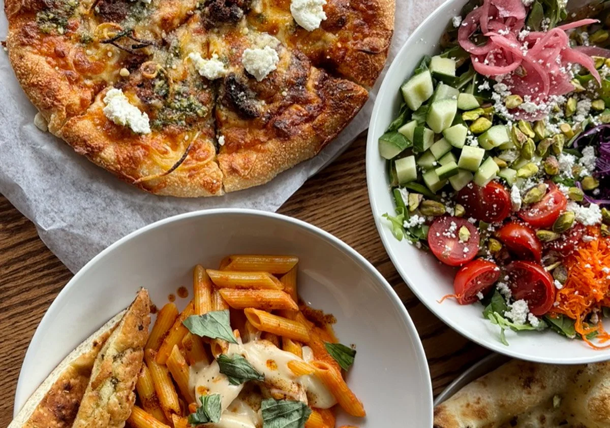 A pizza with cheese and toppings, a pasta dish with tomato sauce, a salad with cherry tomatoes, cucumbers, pistachios, and pickled onions, and a portion of garlic bread.