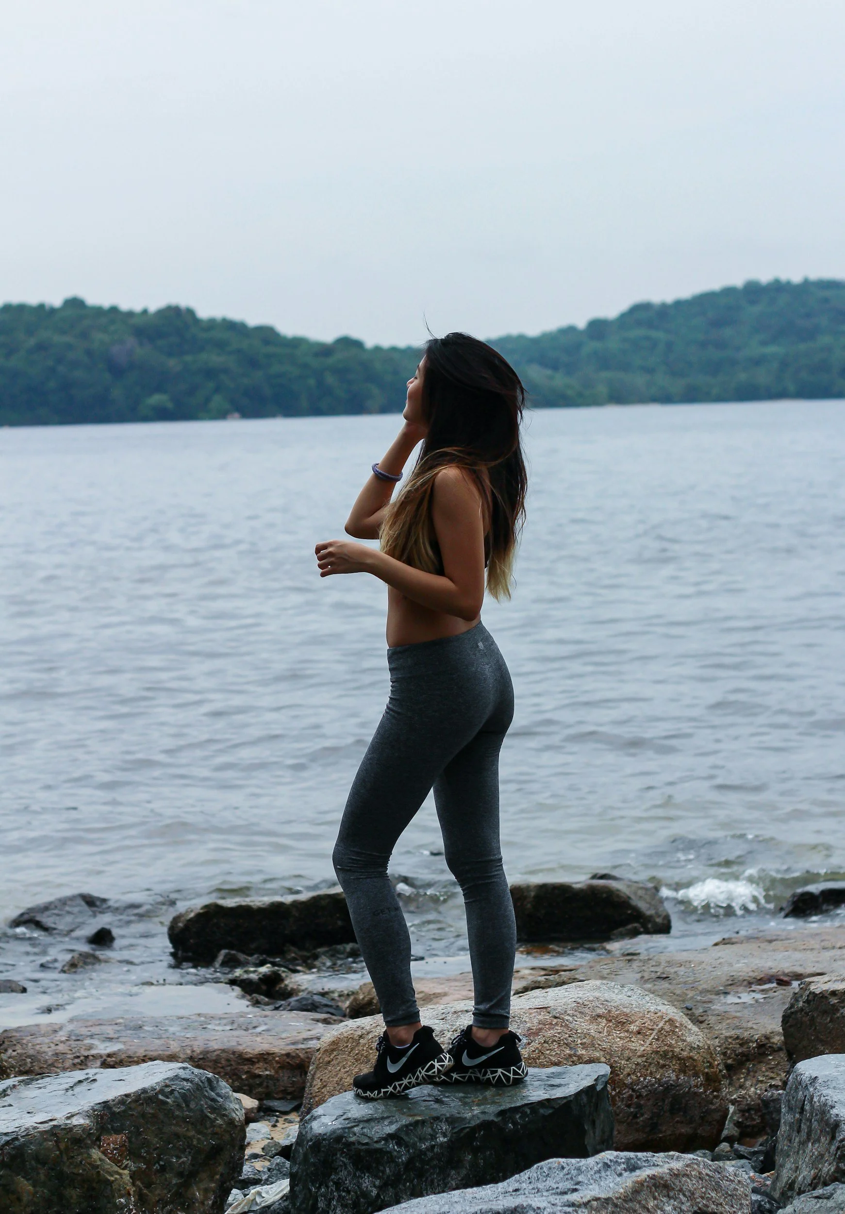 A woman standing on rocks near a lake, wearing gray leggings and black Nike sneakers, with her hand on her head and long hair, overlooking the water and distant island or shoreline.