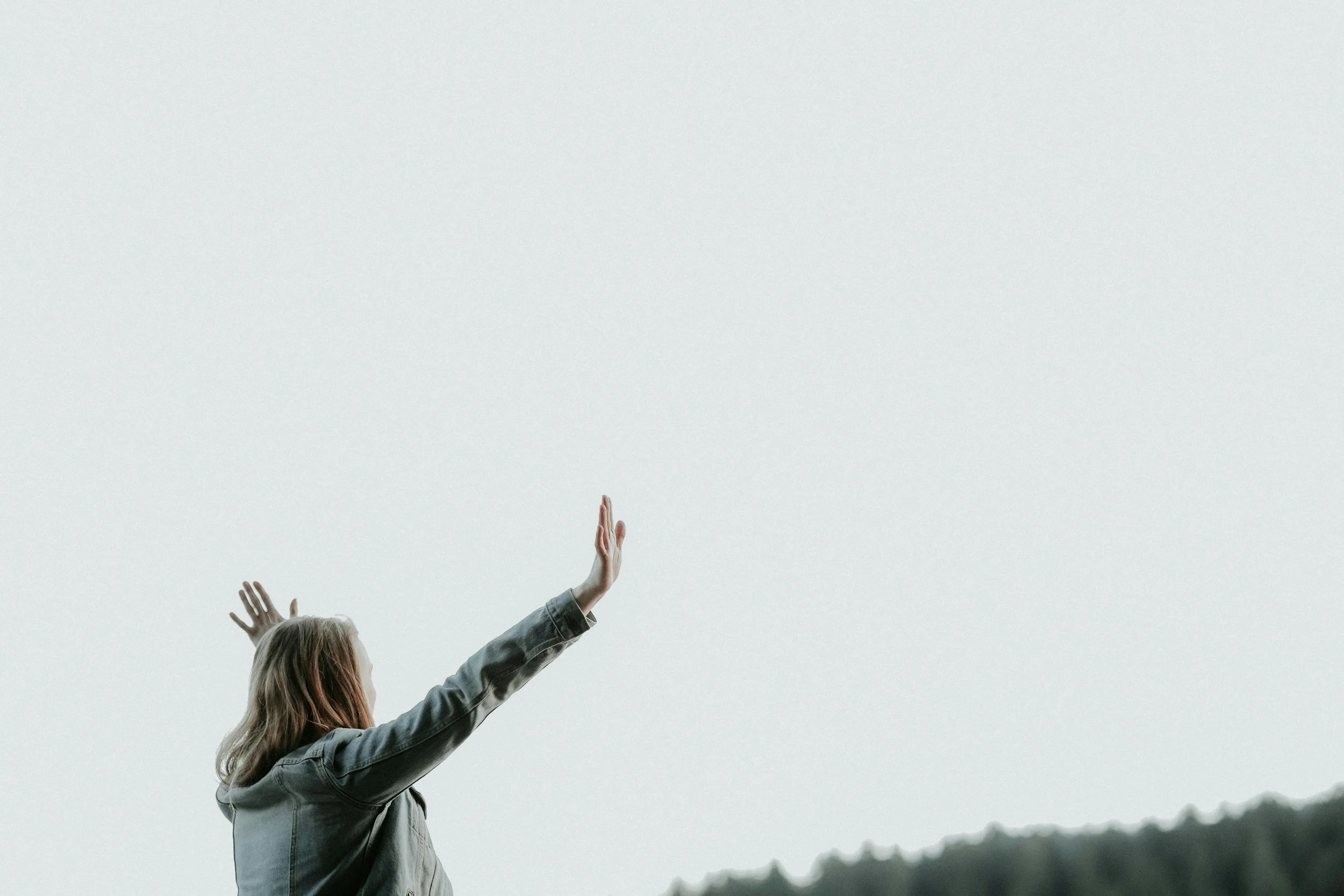 A woman facing the sky with arms raised and palms open, standing outdoors with a hill in the background.