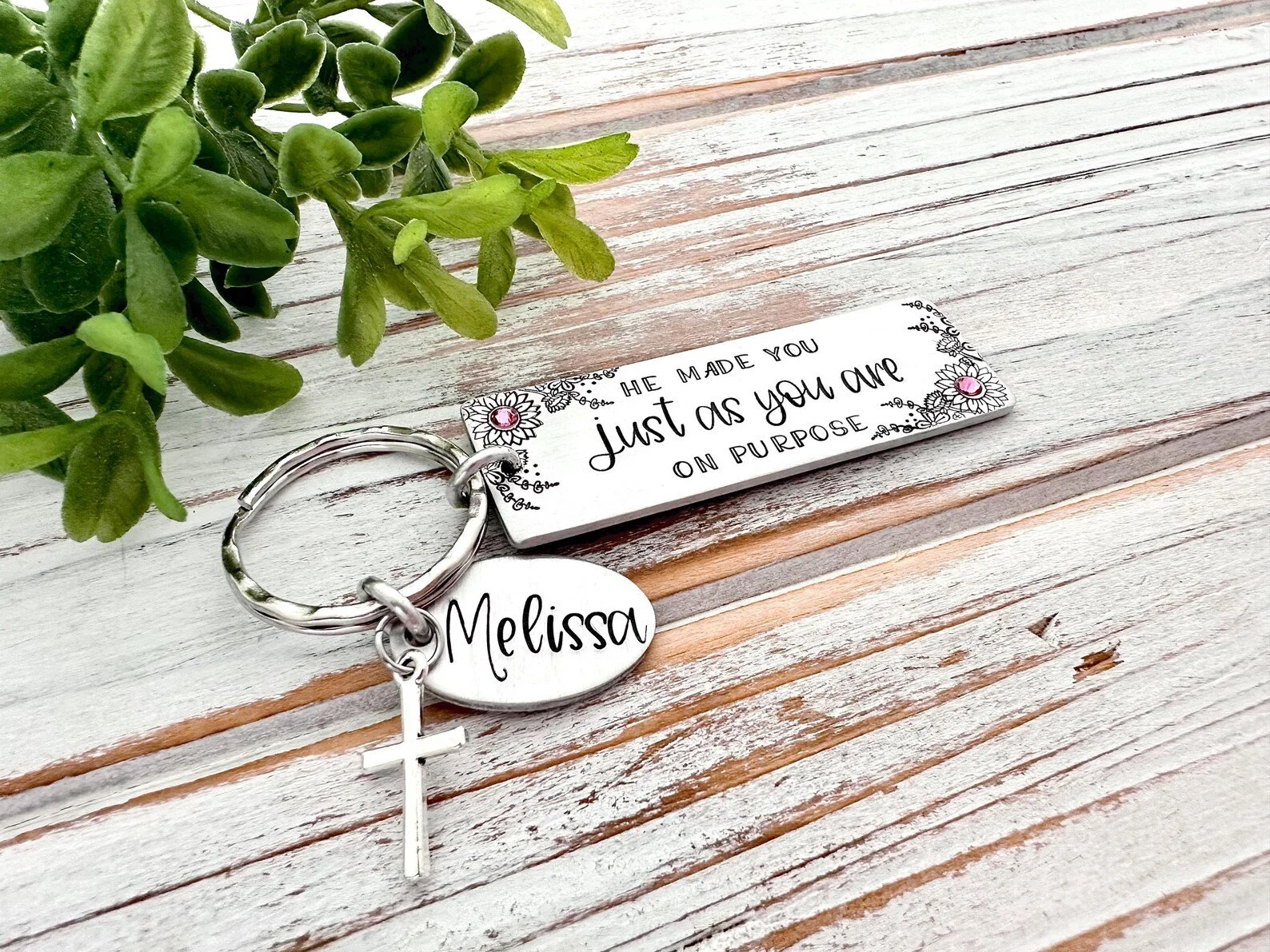 A silver keychain with a personalized oval tag reading 'Melissa,' a white cross, and a rectangular tag with the quote, 'He made you just as you are on purpose,' placed on a rustic white wooden surface with a green leafy plant nearby.