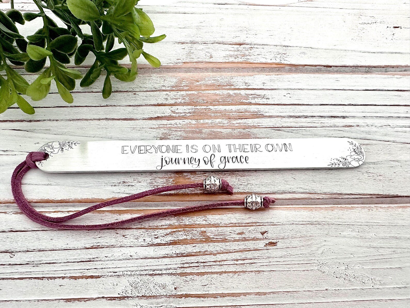 A white wooden bookmark with the written phrase "Everyone is on their own journey of grace" and a purple cord with metal beads attached, placed on a light wooden surface with a green plant in the corner.